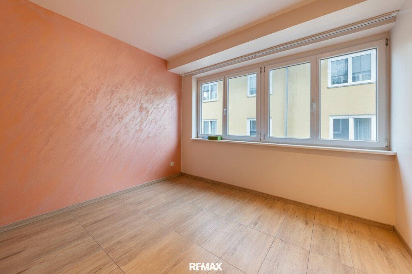 Helles Zimmer mit großen Fenstern, gefliestem Boden und einer Wand in Terrakotta-Optik.