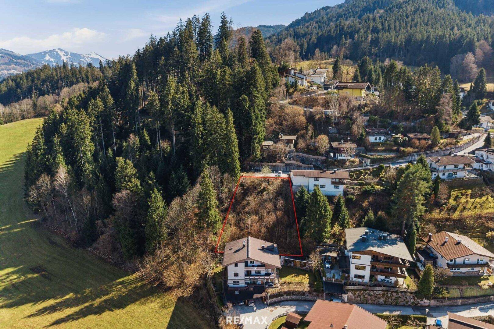 Panoramablick auf ein Baugrundstück in einer grünen Bergregion mit angrenzenden Wohnhäusern.