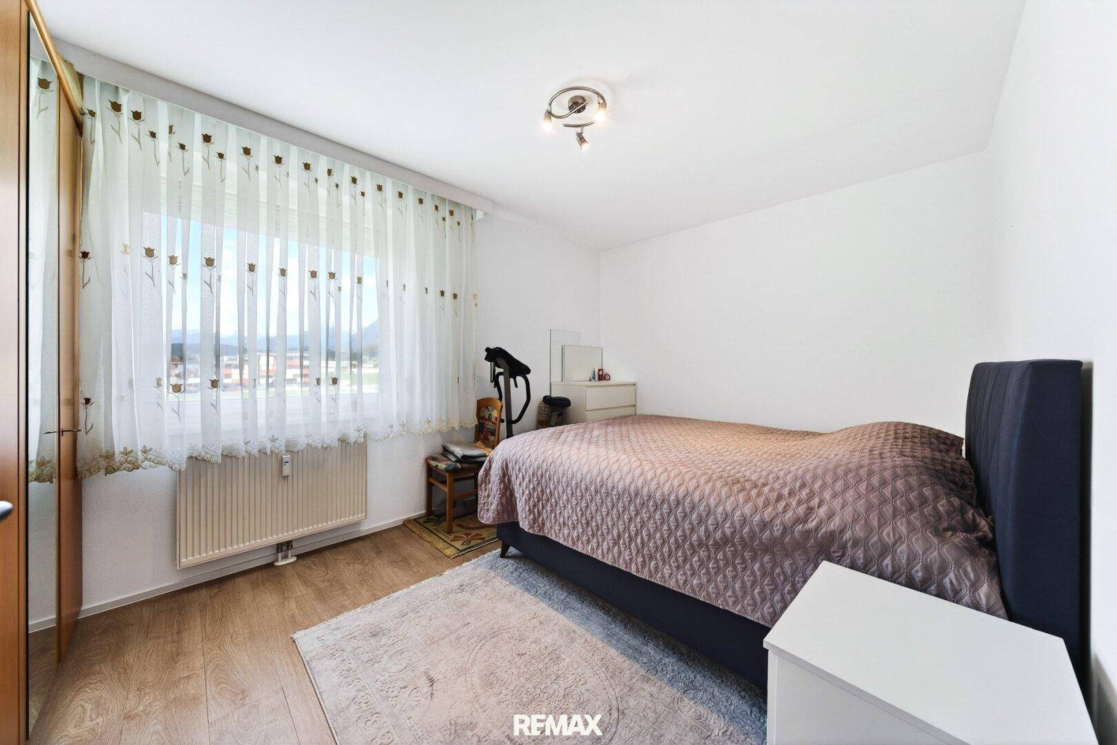 Schlafzimmer mit Doppelbett, Holzschrank und großem Fenster, das viel Tageslicht hereinlässt.