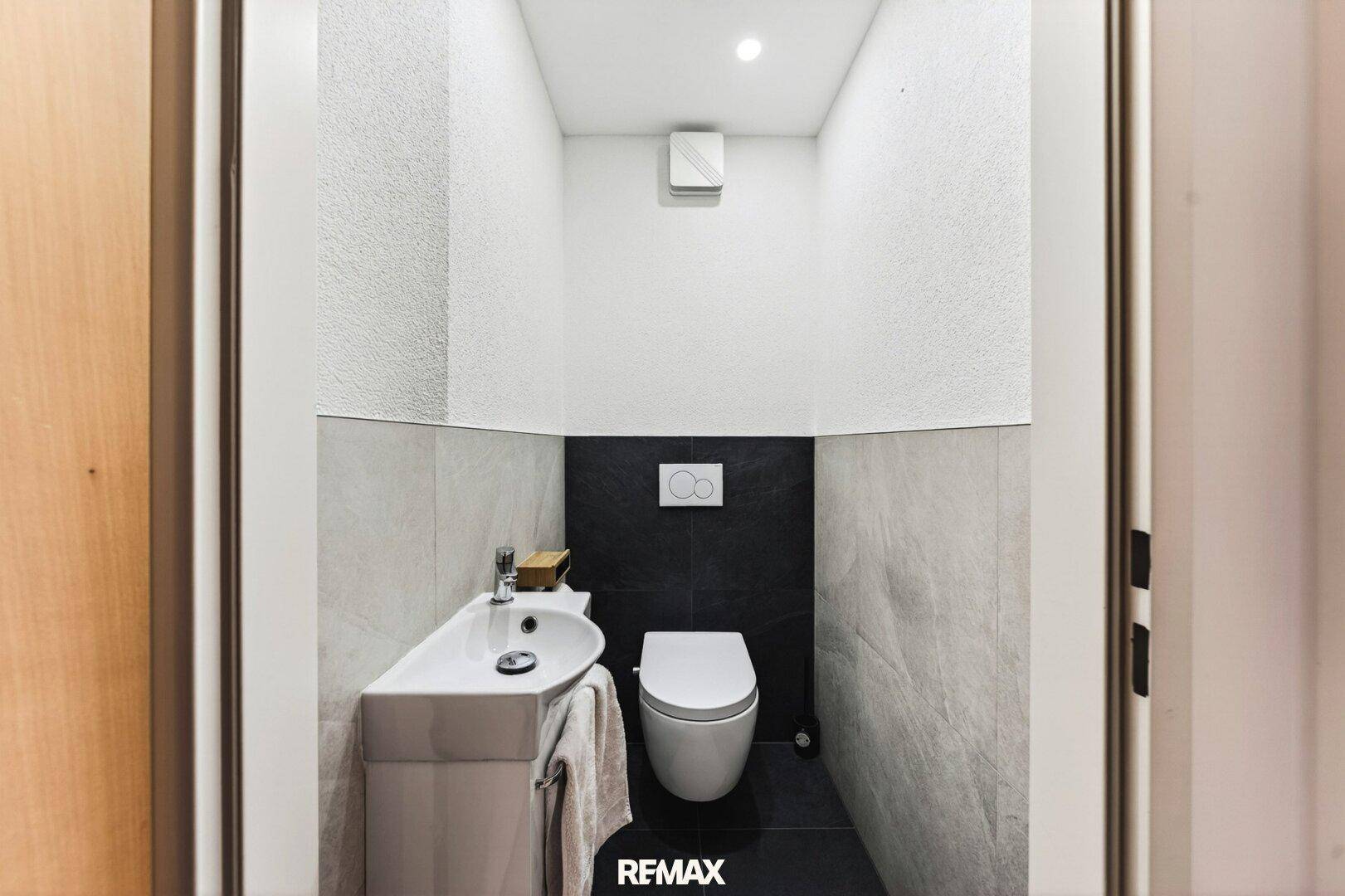 Modernes Gäste-WC mit Wand-WC, kleinem Waschbecken und dunklen Fliesen im unteren Bereich.