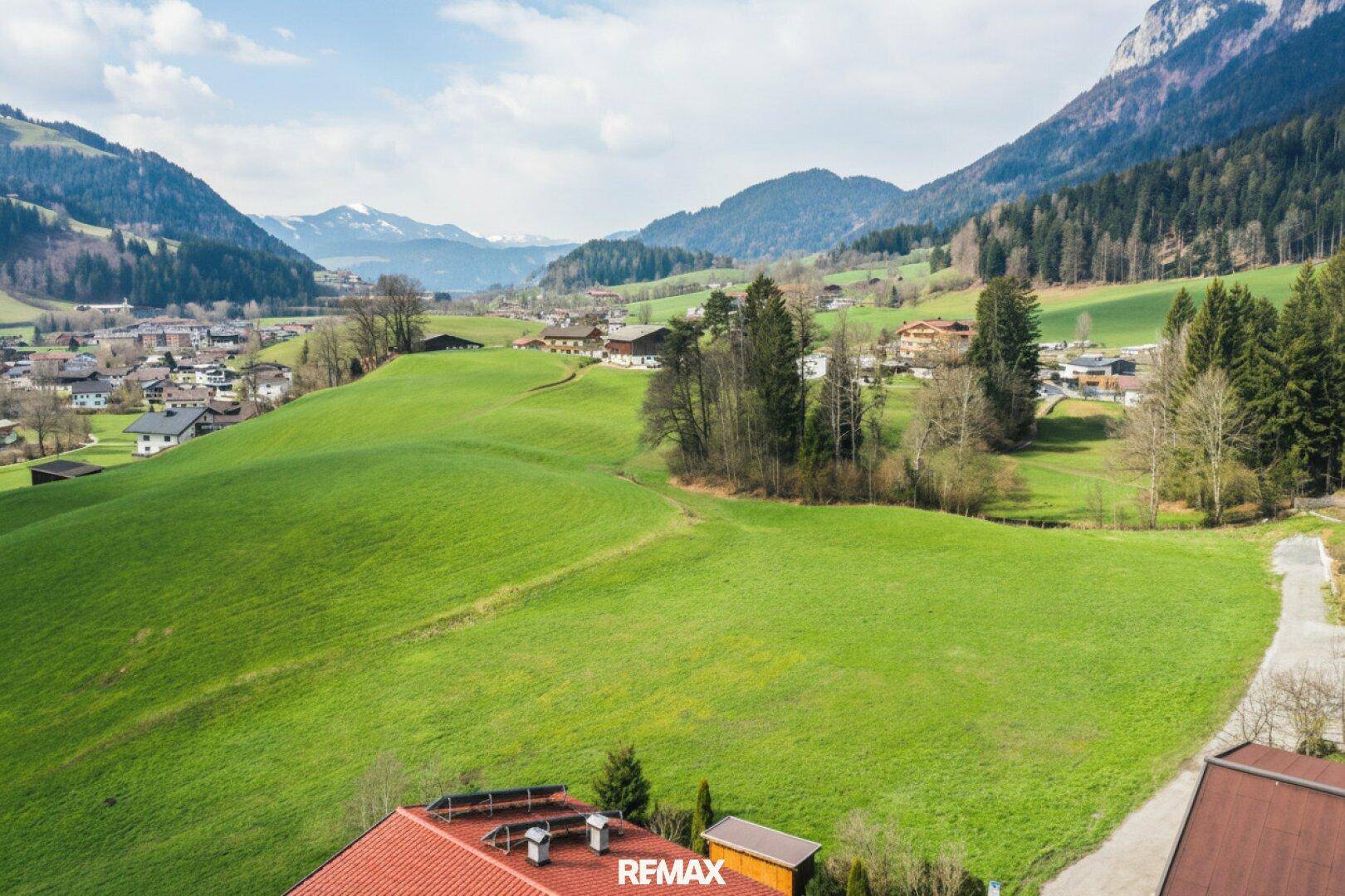 Panoramablick über sanfte grüne Hügel, ein idyllisches Dorf und die umliegende Berglandschaft.