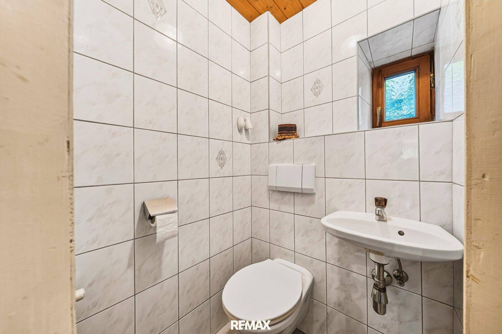 Kleines WC mit weißen Fliesen, Toilette, Waschbecken und einem Fenster für natürliche Belüftung.