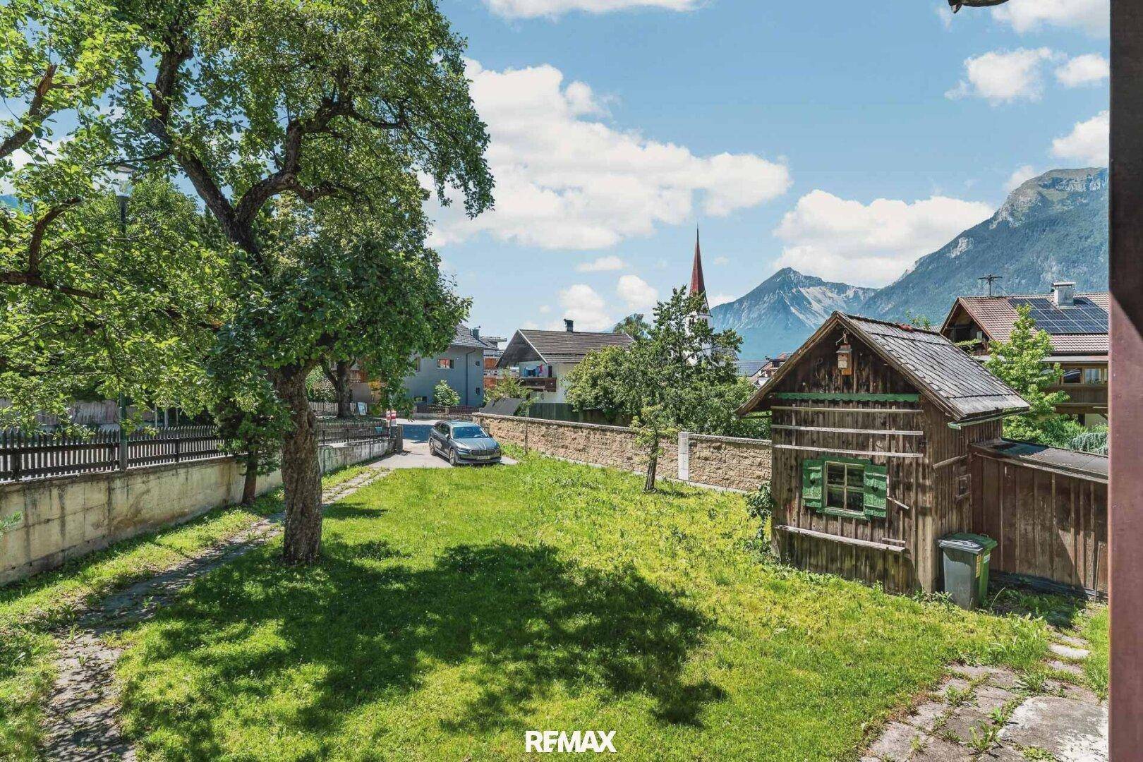 Grüner Gartenbereich mit altem Baum, Holzhütte und Blick auf die Berge im Hintergrund.