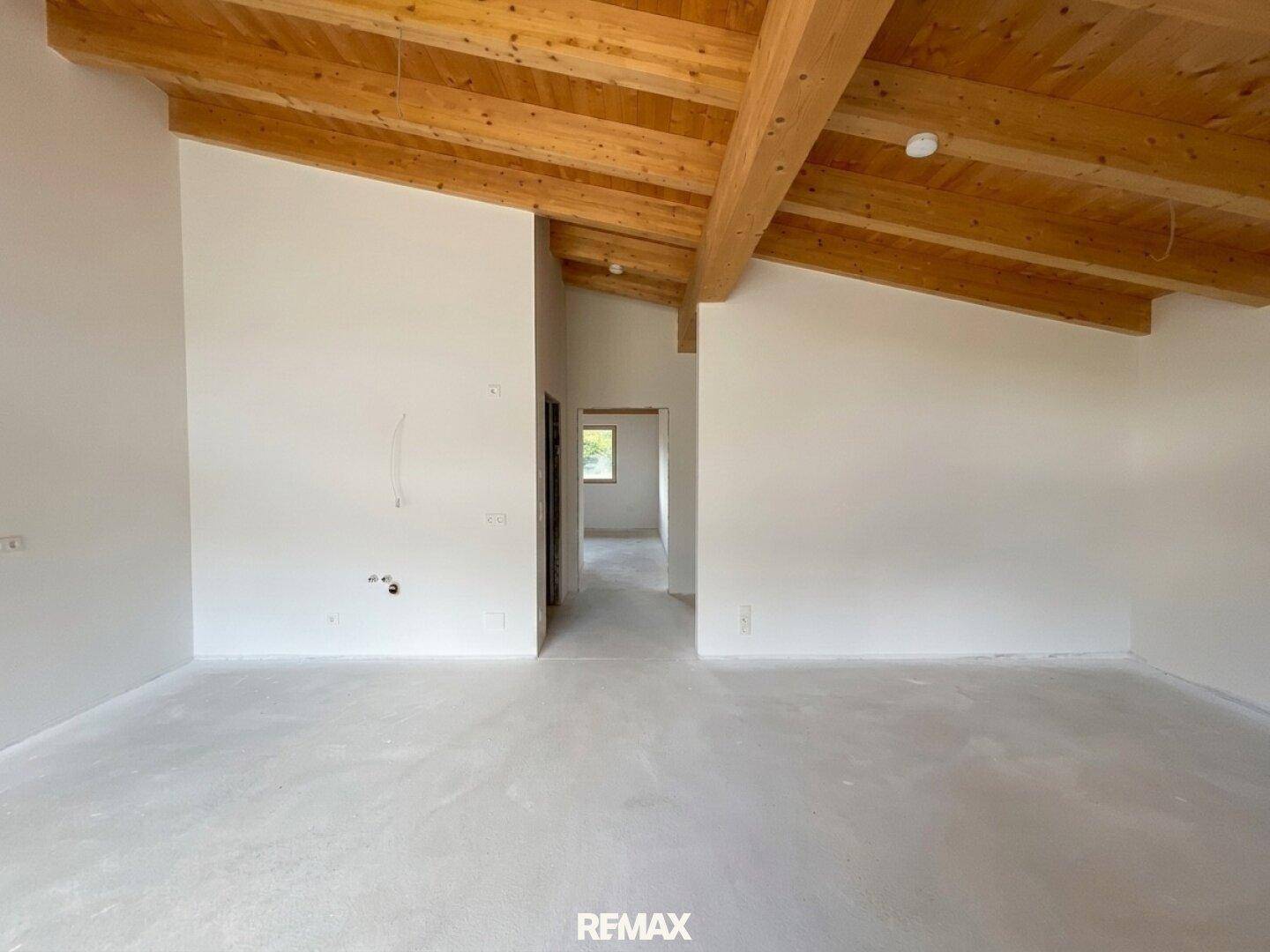 Offener Innenraum mit markanten Holzdeckenbalken und Betonboden, führt zu weiteren Zimmern im modernen Neubau.