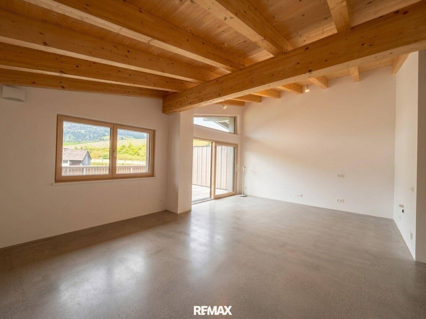Heller Wohnraum mit rustikalen Holzdeckenbalken und modernem Betonboden, mit großen Fenstern für viel Tageslicht.