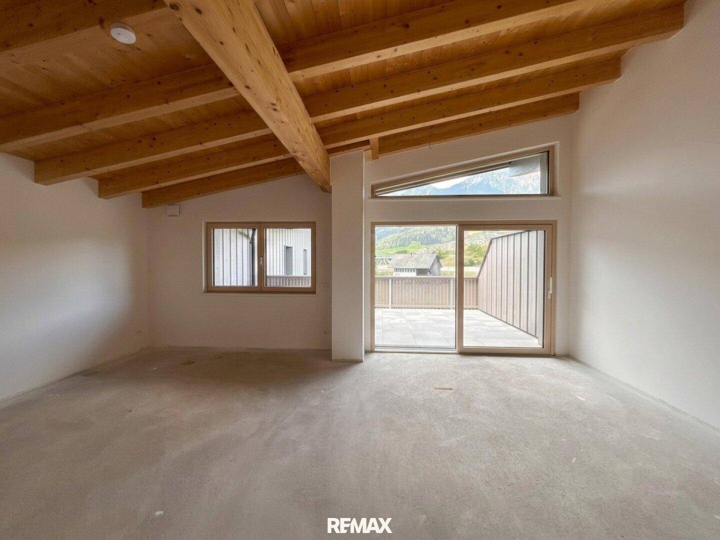 Geräumiger Wohnbereich mit sichtbaren Holzdeckenbalken und Betonboden, bietet direkten Zugang zur Terrasse mit Bergblick.