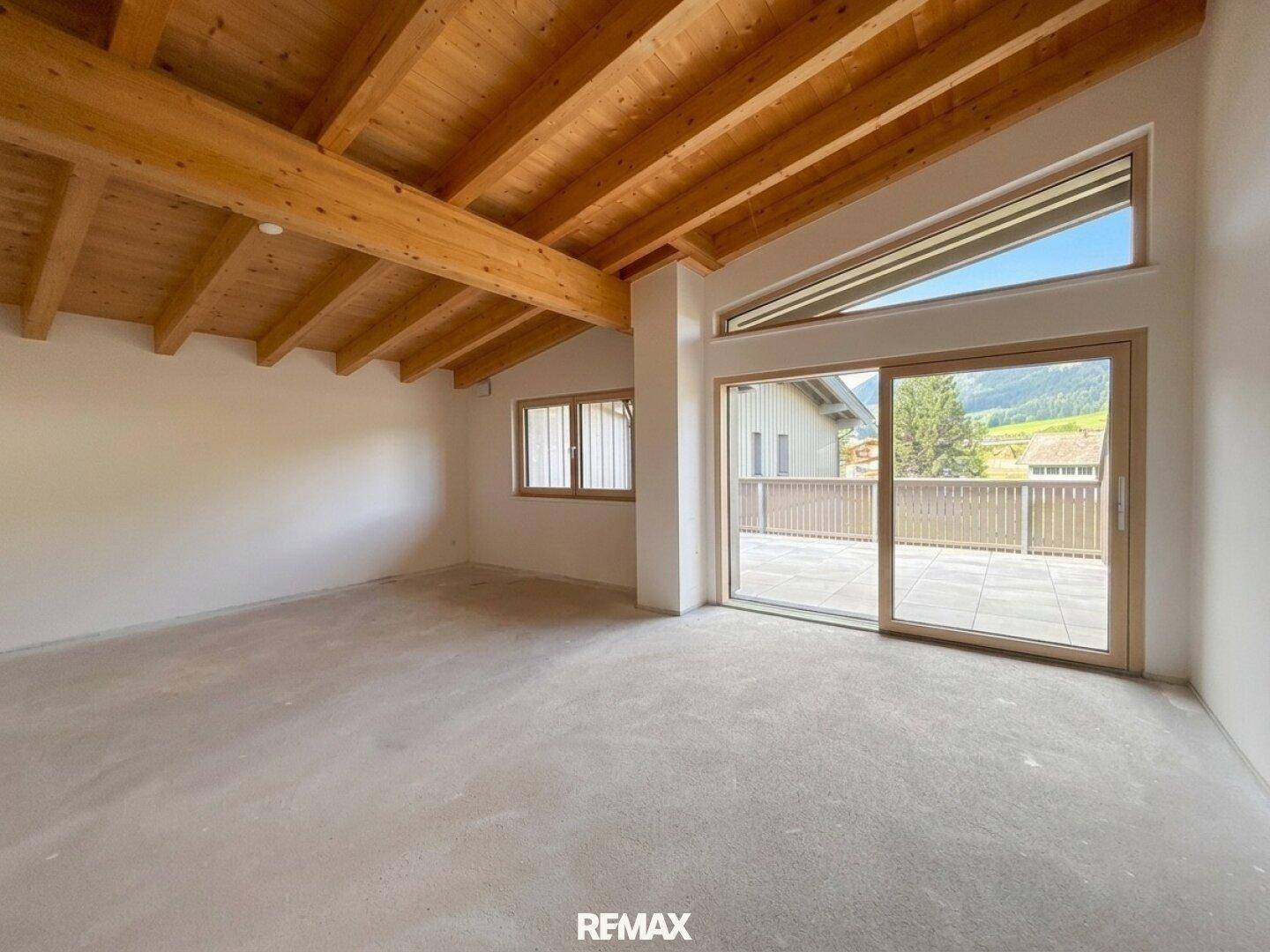 Heller, unmöblierter Wohnraum mit sichtbaren Holzdeckenbalken, Betonboden und Zugang zu einer Terrasse mit Bergblick.