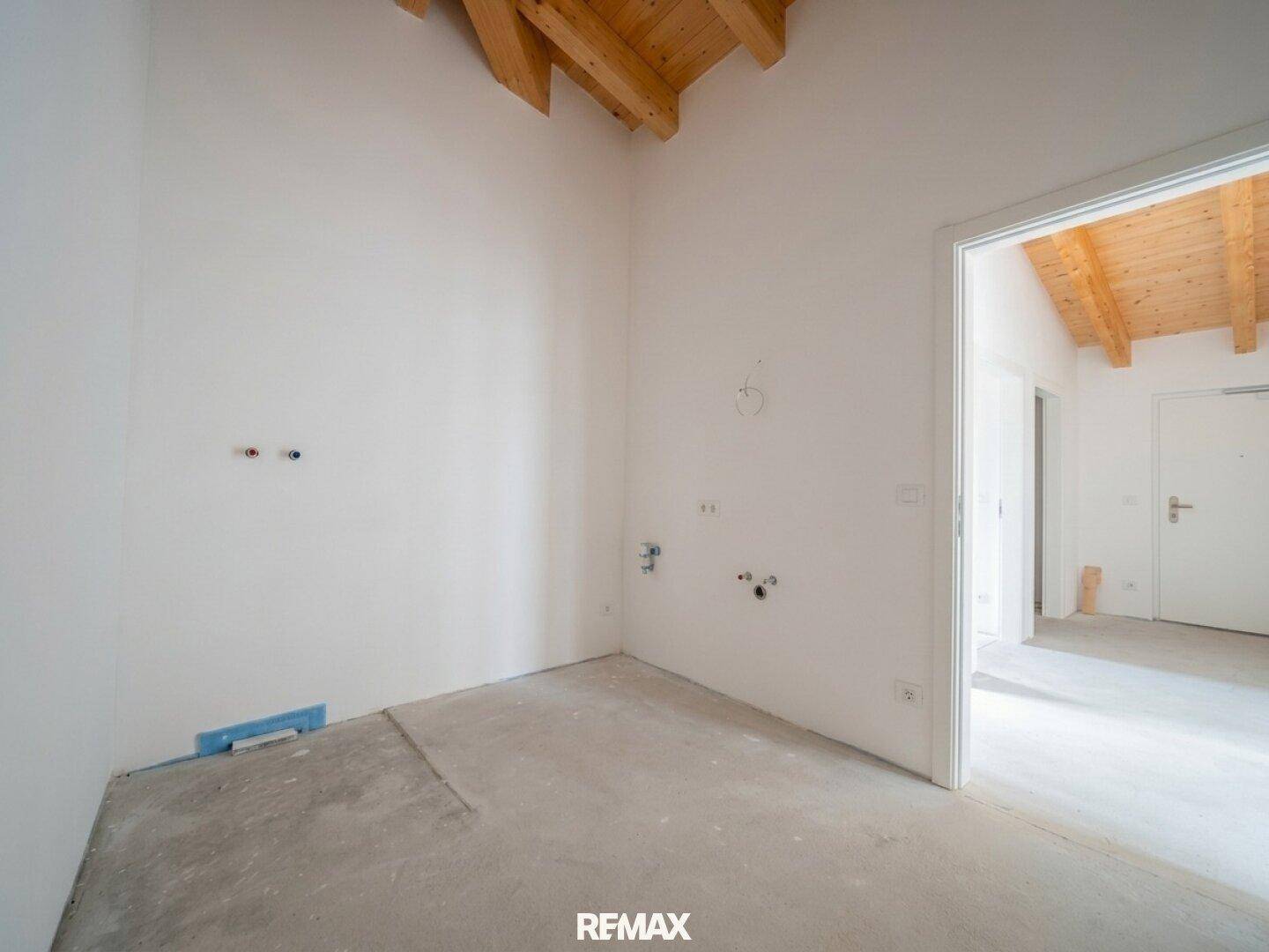 Unmöblierte Küche mit sichtbaren Anschlüssen, Holzdeckenbalken und Betonboden, bereit für die individuelle Gestaltung.