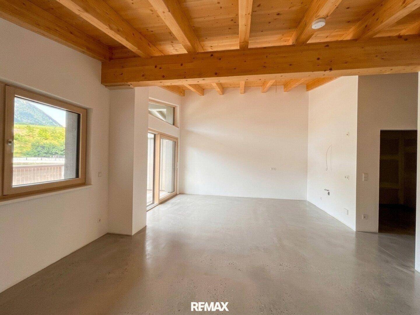 Lichtdurchfluteter Wohnbereich mit sichtbaren Holzdeckenbalken und einem schlichten Betonboden, bietet viel Gestaltungspotenzial.