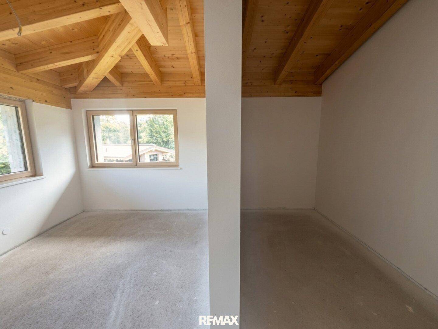 Heller, unmöblierter Raum mit Holzbalkendecke und Betonboden, durch eine Wand geteilt, mit Fenster.