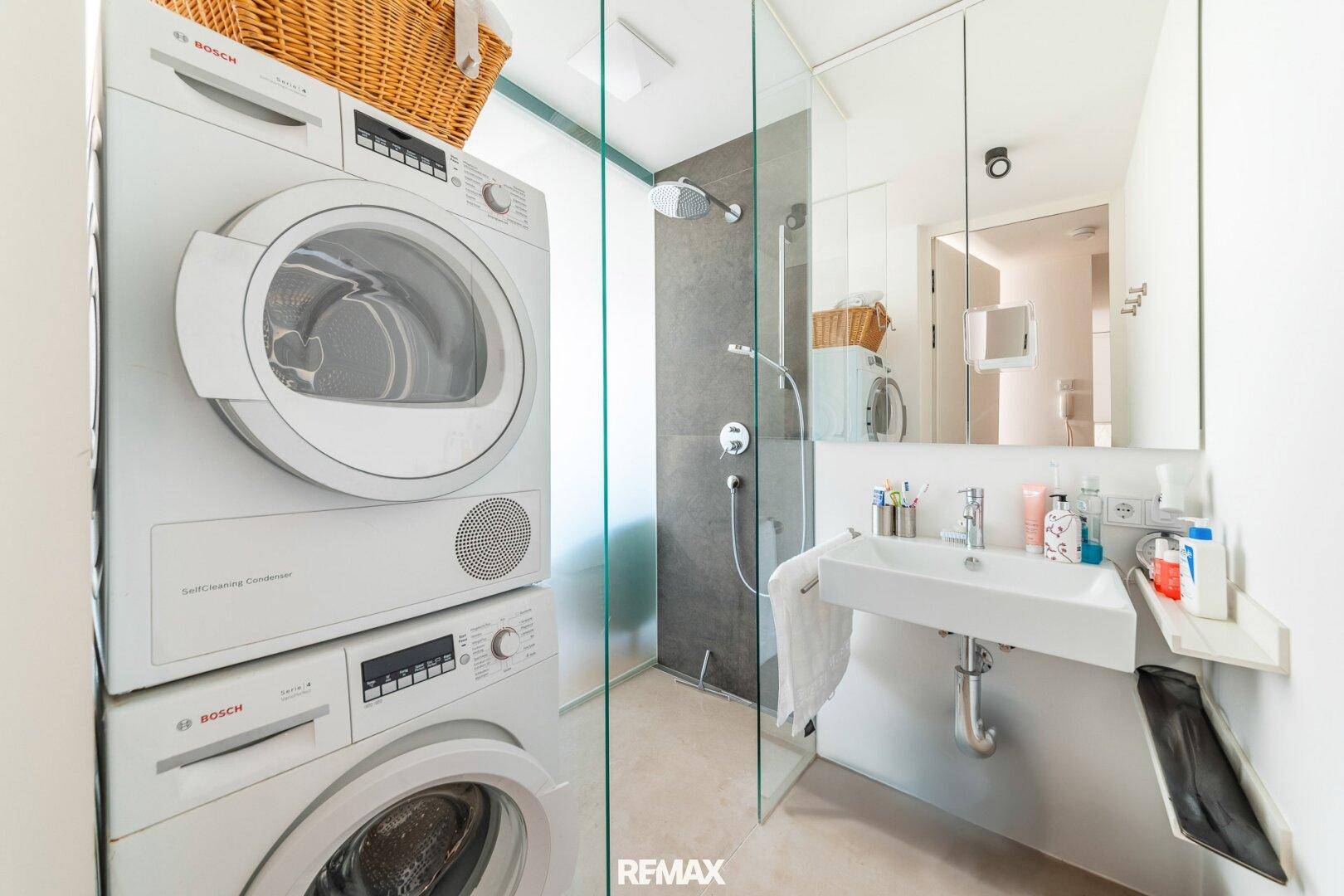 Modernes Badezimmer mit Waschmaschine, Trockner und einer begehbaren Dusche mit Glaswand.