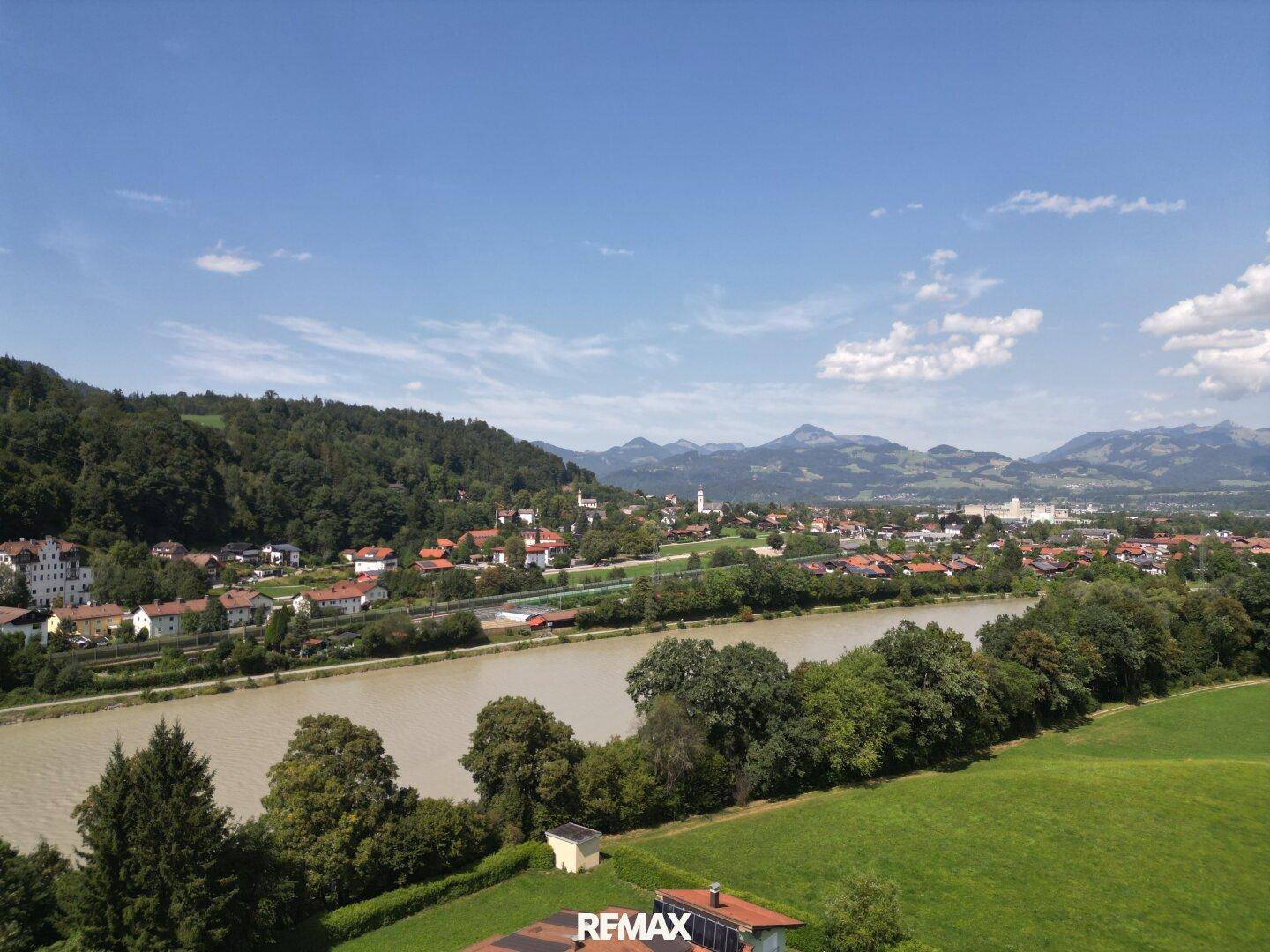 Weitläufige Landschaftsaufnahme mit einem Fluss, grünen Wiesen, einer Stadt und majestätischen Bergen im Hintergrund.