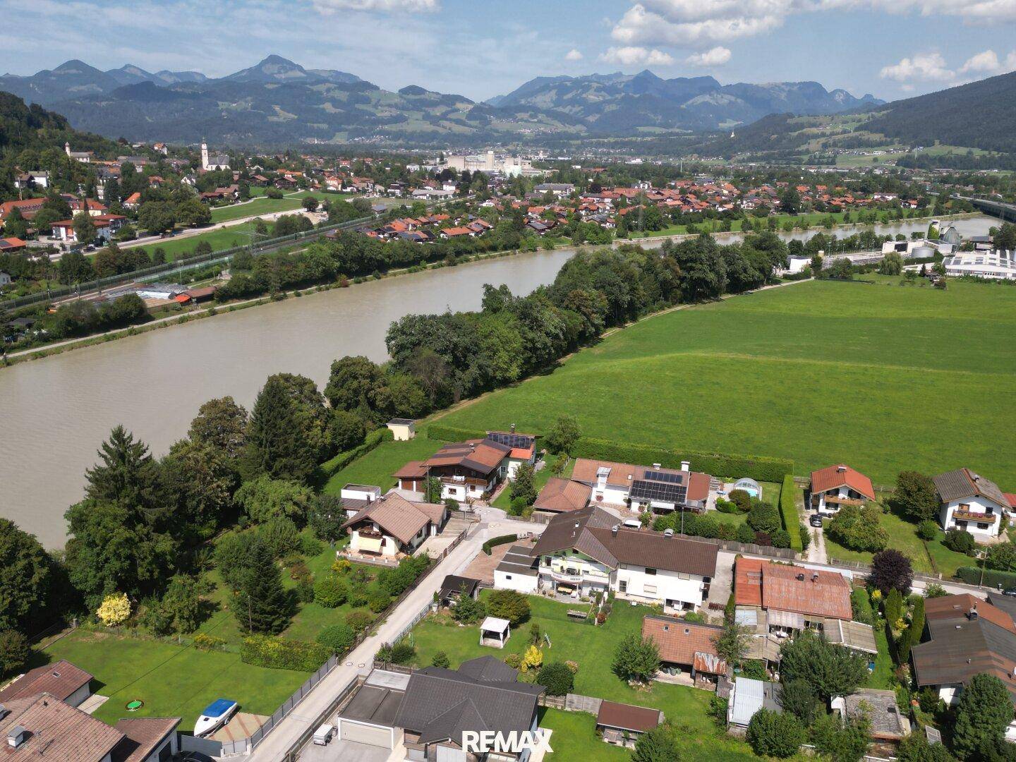 Panoramablick aus der Luft auf ein Wohngebiet entlang eines Flusses mit grünen Feldern und Bergen.