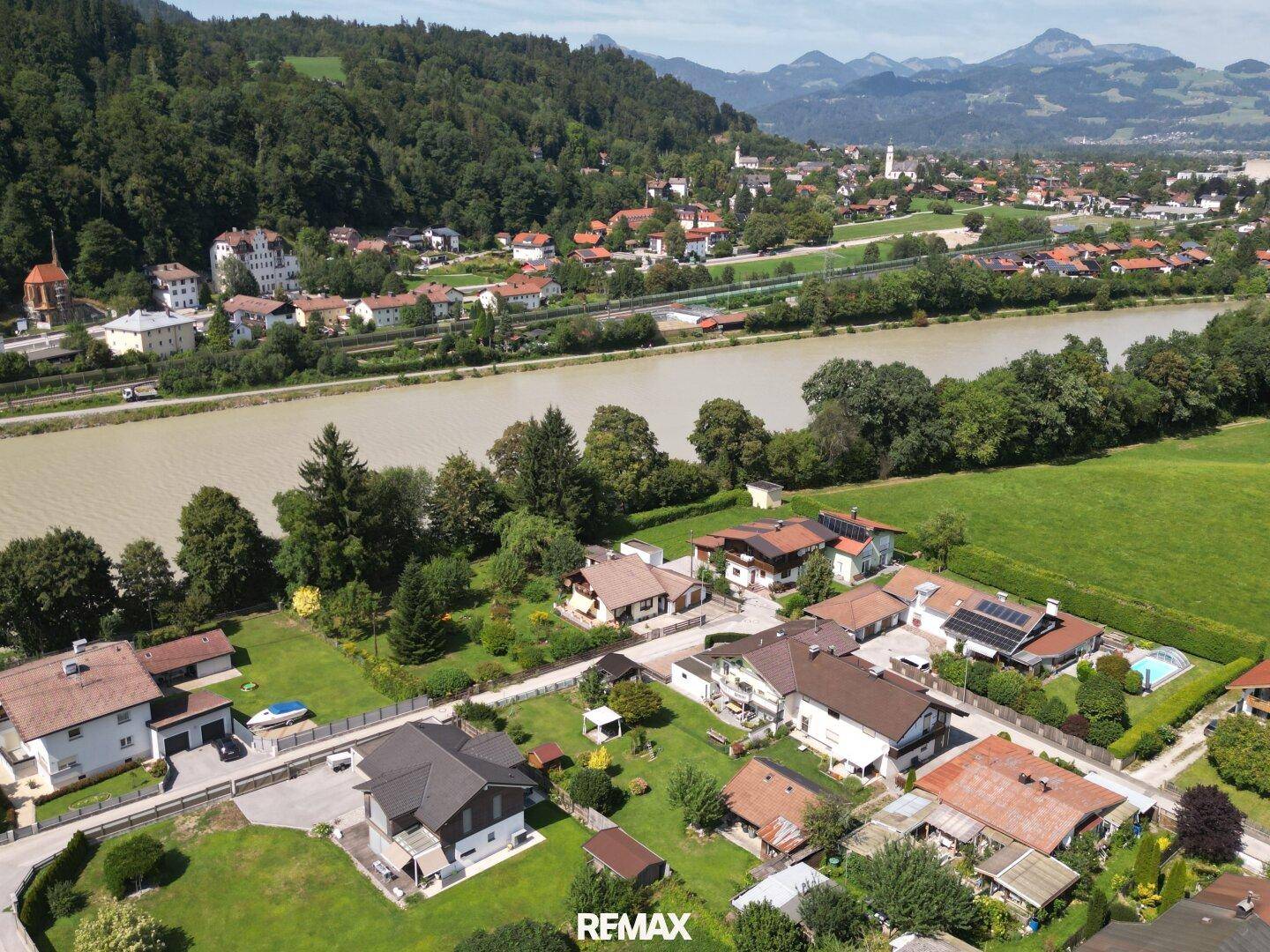 Panoramablick aus der Luft auf ein Wohngebiet entlang eines Flusses mit grünen Feldern und Bergen.