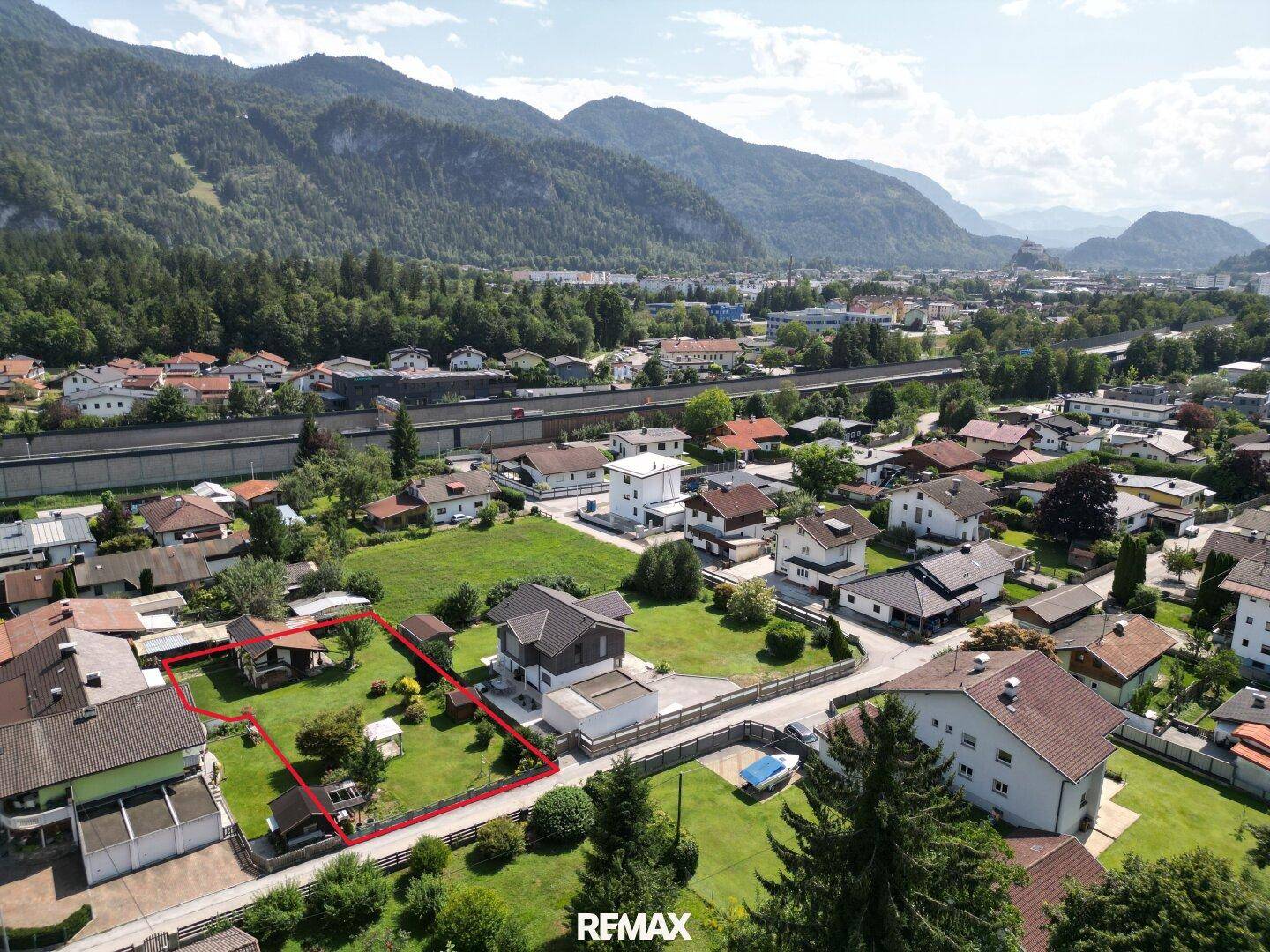Weitläufige Luftaufnahme eines Wohngebiets mit Bergen im Hintergrund und einem rot markierten Grundstück.
