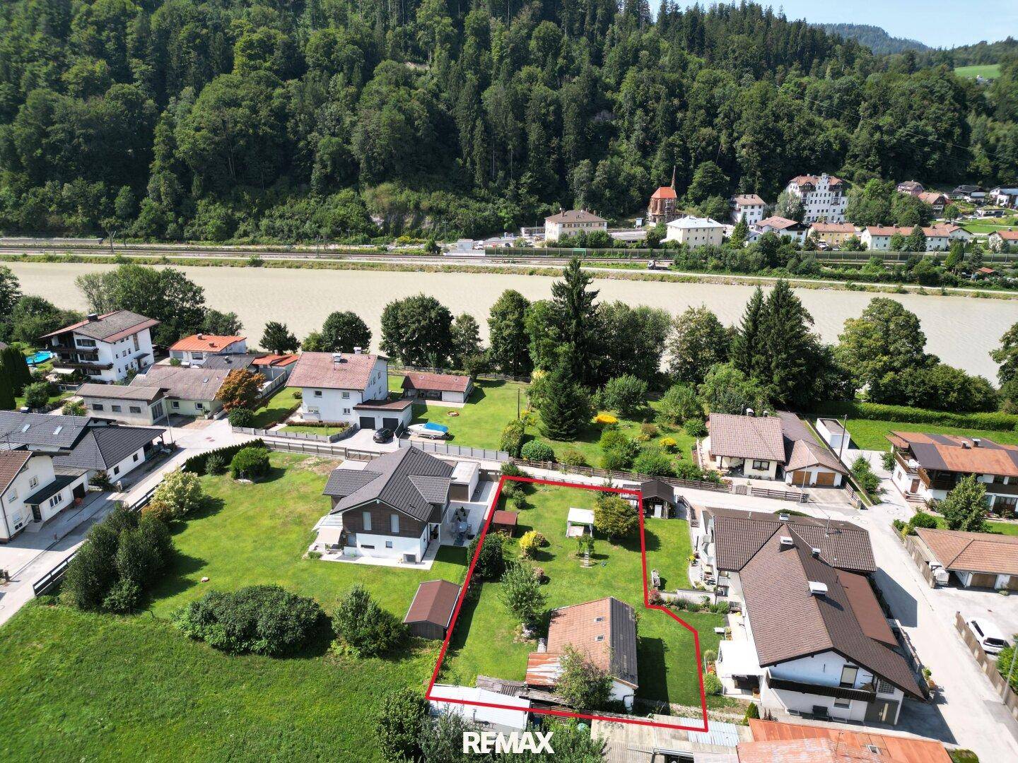 Luftaufnahme eines Wohngebiets mit Fluss, Bergen und einem rot umrandeten Grundstück in ländlicher Umgebung.