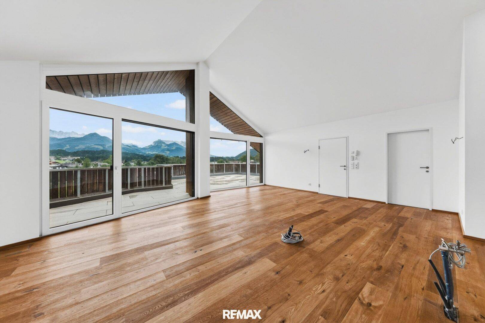 Heller, leerer Wohnraum mit Holzboden, hohen Decken und großen Fenstern mit Bergblick.