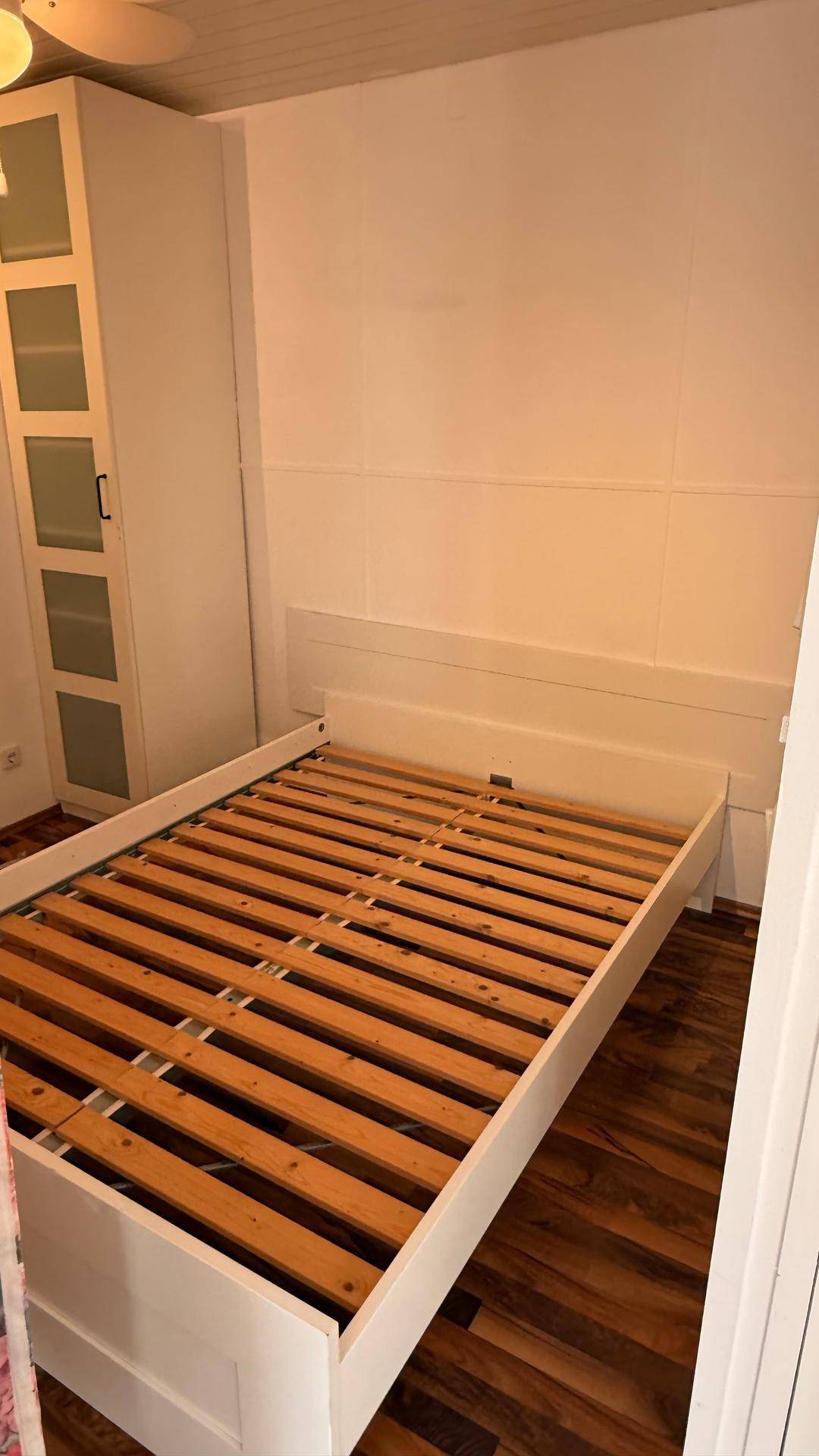 Schlafzimmer mit einem Bettgestell, Lattenrost und einem hohen Schrank für Kleidung.