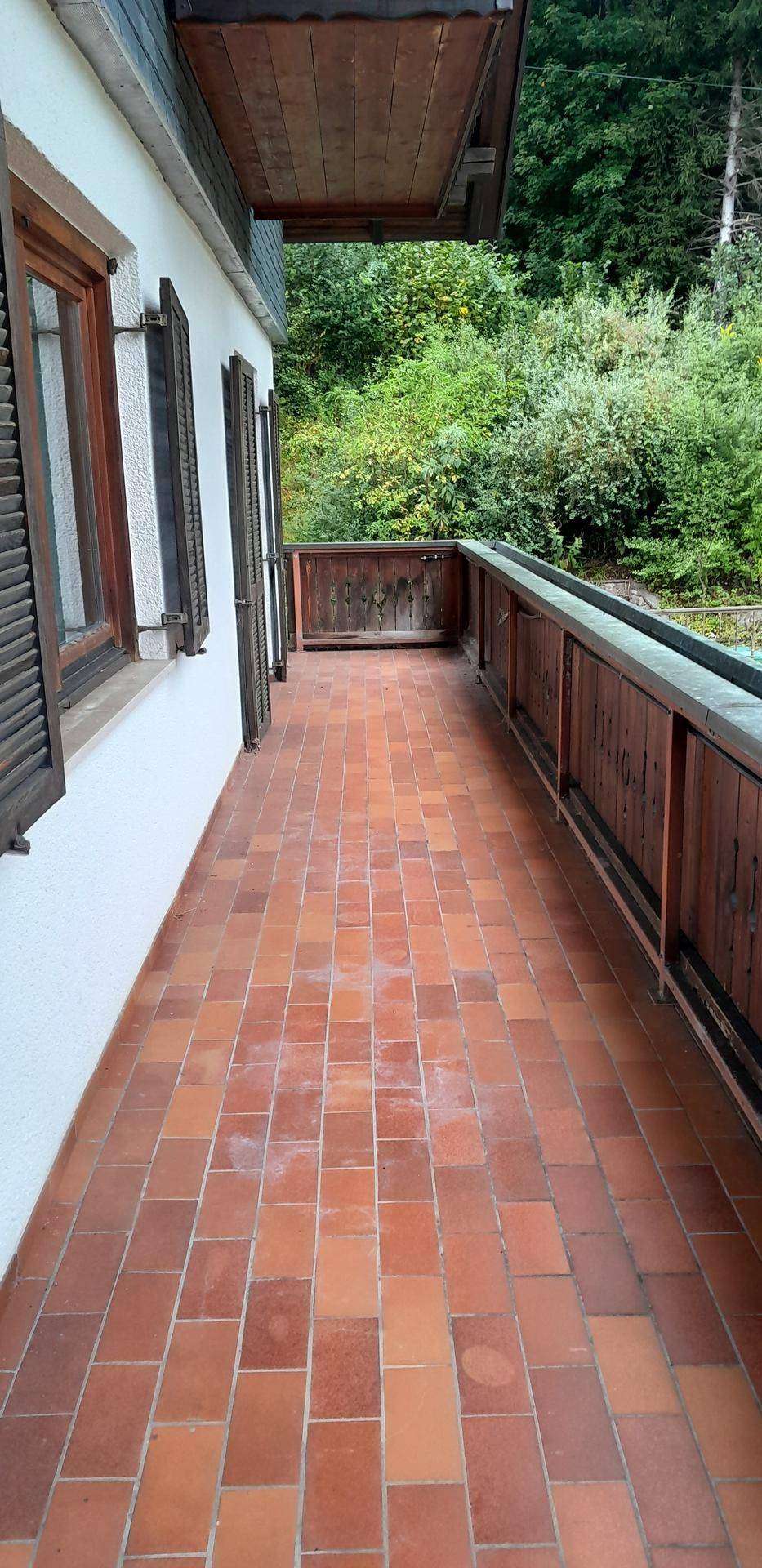 Langer Balkon mit Terrakottafliesen und Holzgeländer, umgeben von viel Grün.