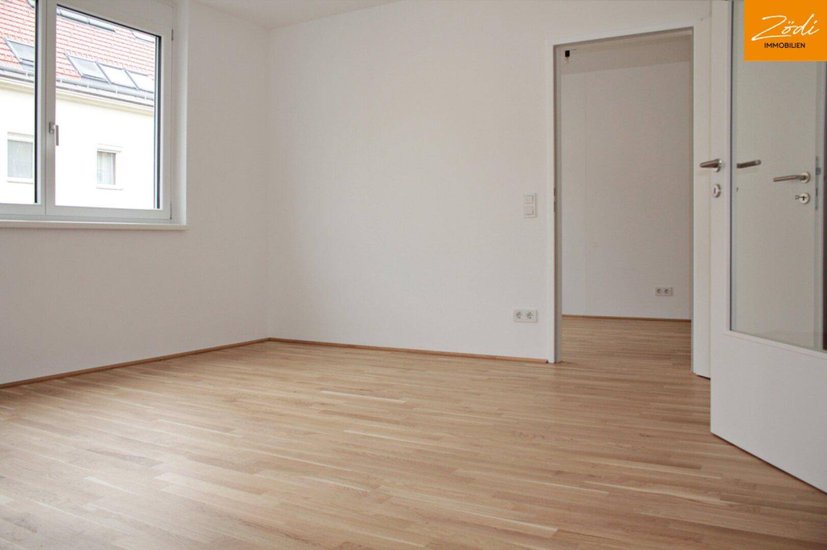 Leeres Zimmer mit hellem Parkettboden und Fenster mit Blick auf die Nachbarschaft.