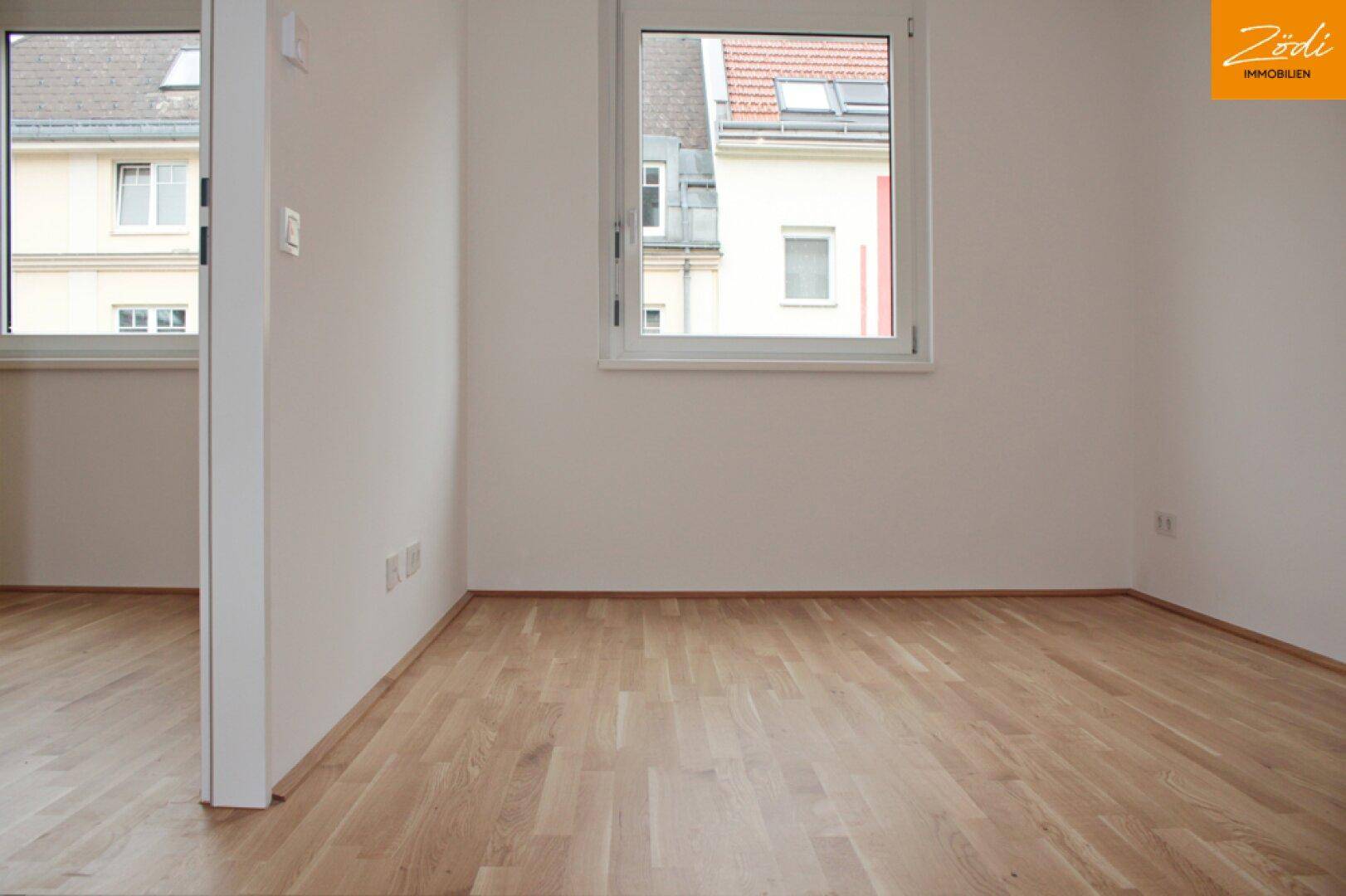 Helles, leeres Zimmer mit Parkettboden und zwei Fenstern für viel Tageslicht.