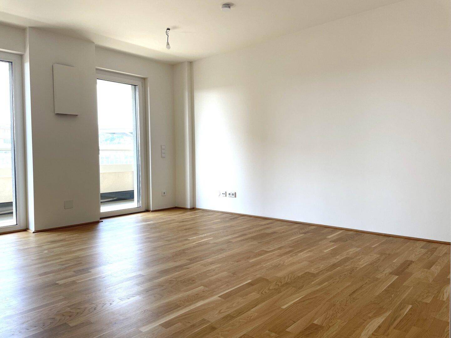 Leeres Wohnzimmer mit hellem Parkettboden und Zugang zu einem Balkon.