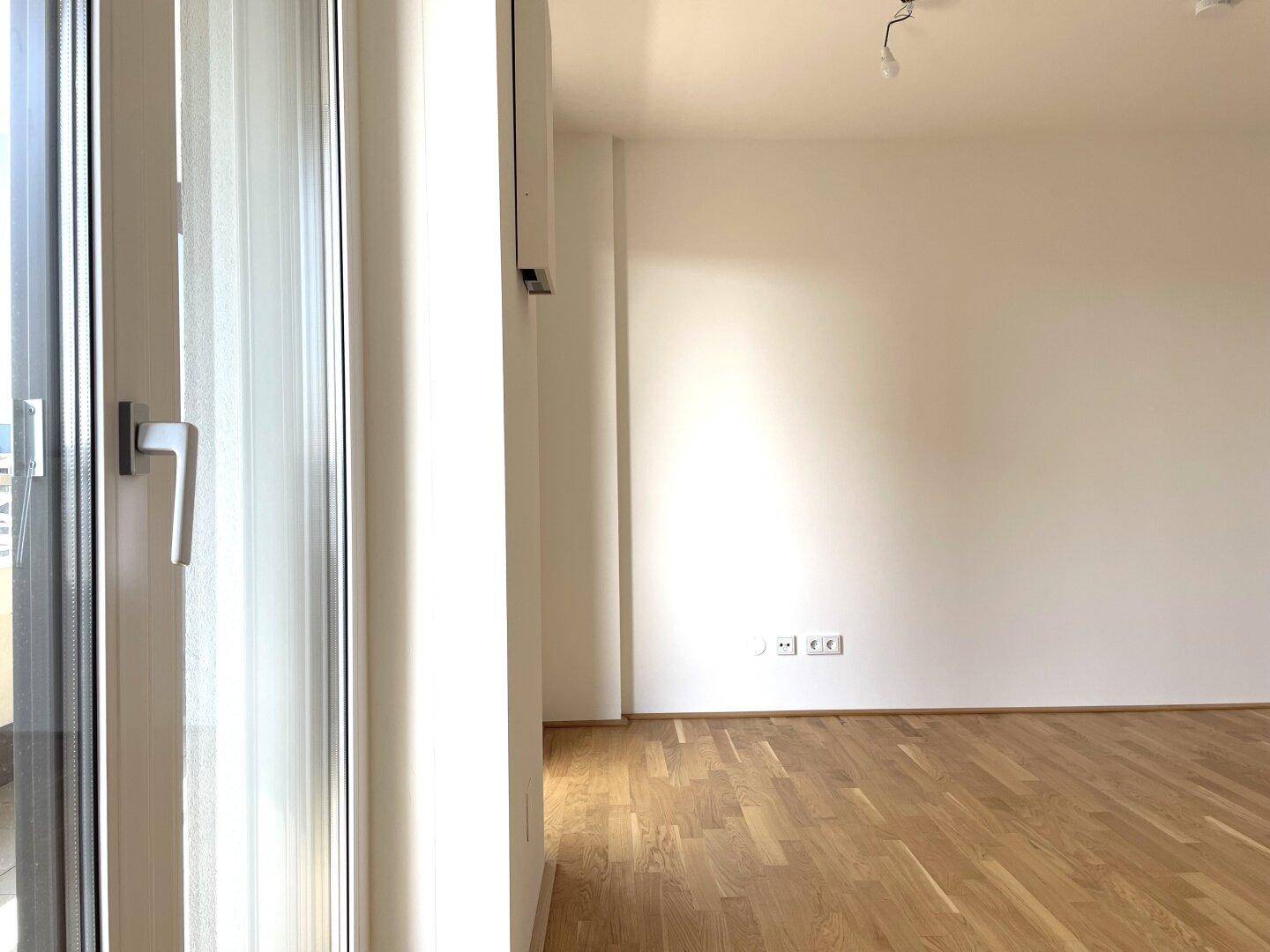 Heller Raum mit Parkettboden und einem großen Fenster, das viel Licht hereinlässt.