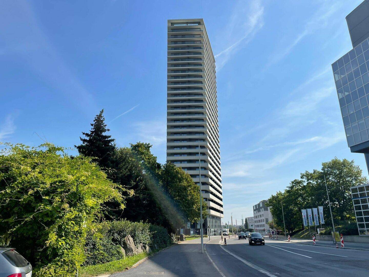 Hohes Wohngebäude mit vielen Balkonen und moderner Architektur, umgeben von Grünflächen und einer Straße.
