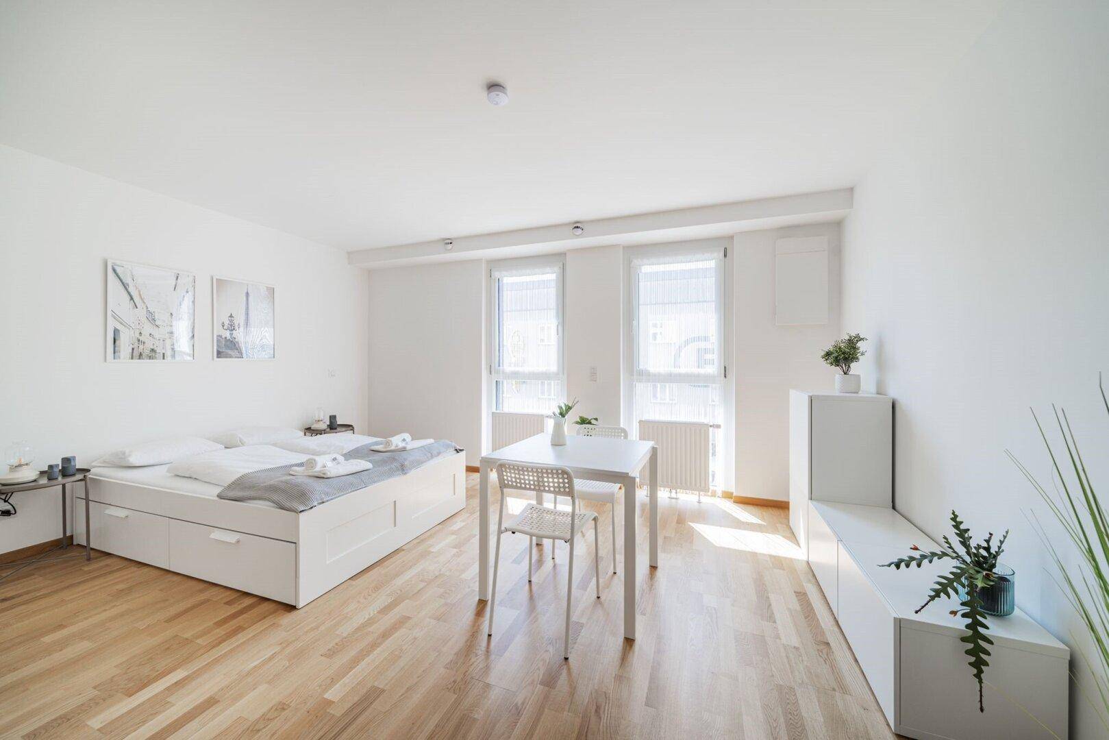 Helles Studio-Apartment mit Doppelbett, Esstisch und großen Fenstern, modern eingerichtet.