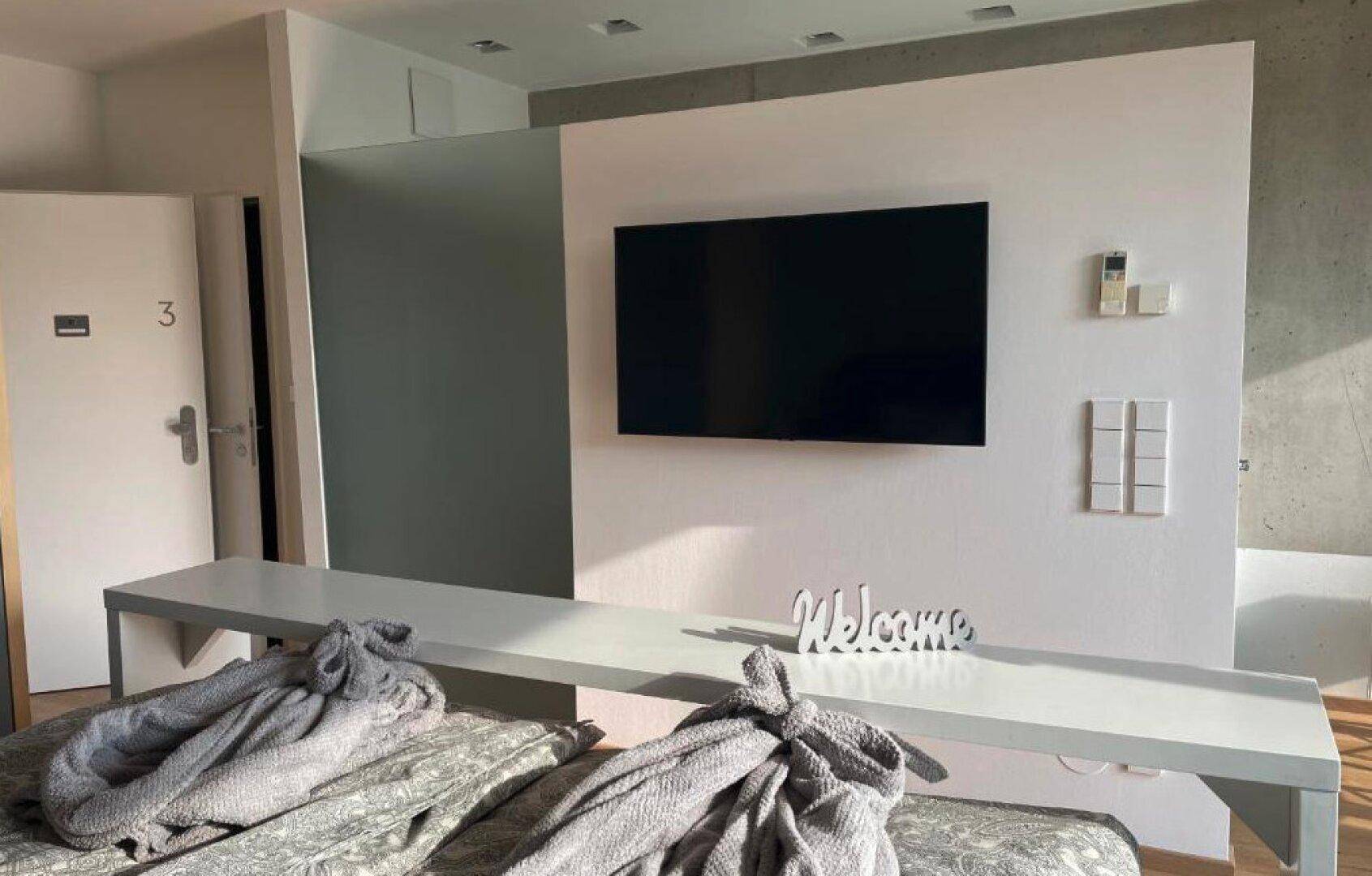 Schlafbereich mit großem Bett, Wandfernseher und "Welcome"-Dekoration.