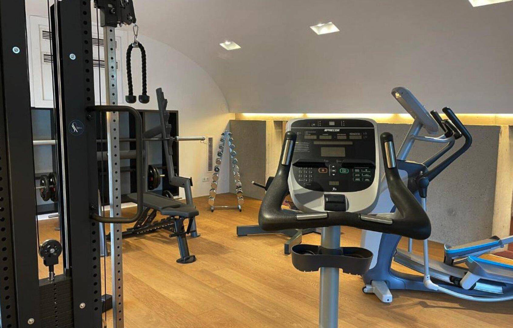 Gut ausgestatteter Fitnessraum mit verschiedenen Trainingsgeräten und hellem Holzfußboden.