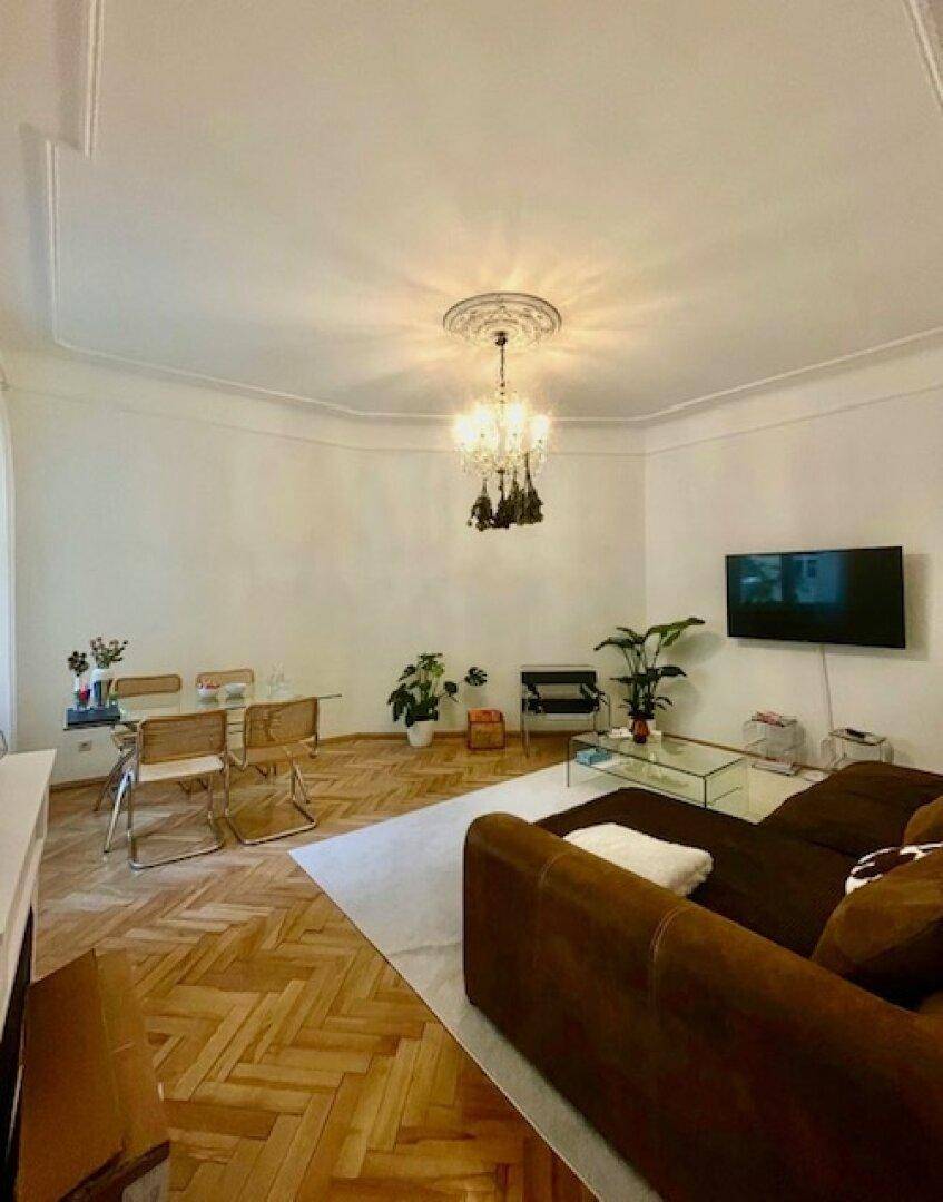Geräumiger Wohn-Essbereich mit Kronleuchter, Fischgrätparkett und Fernseher.