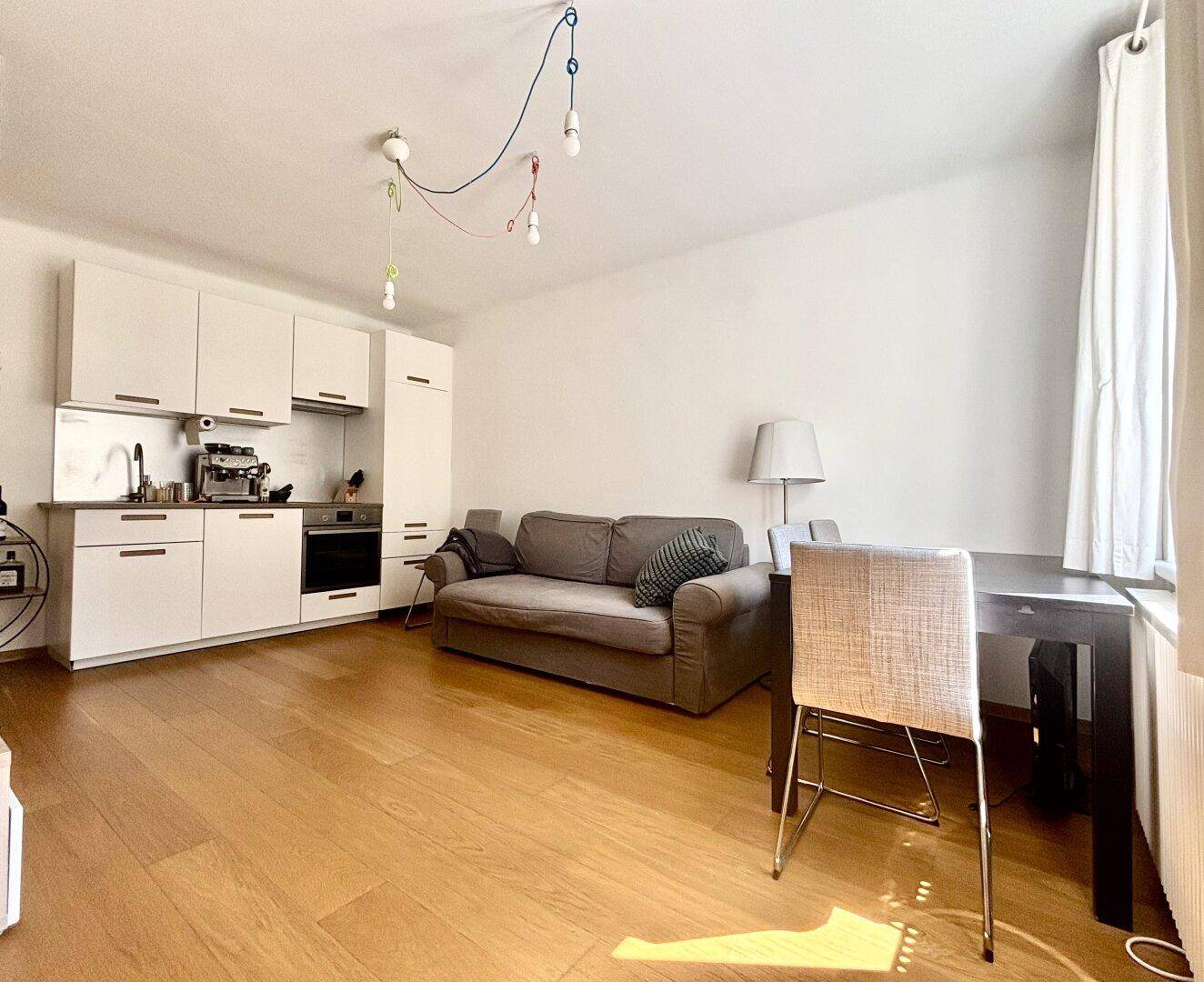 Heller Wohn-Essbereich mit kleiner Küchenzeile, Sofa und Esstisch am Fenster.