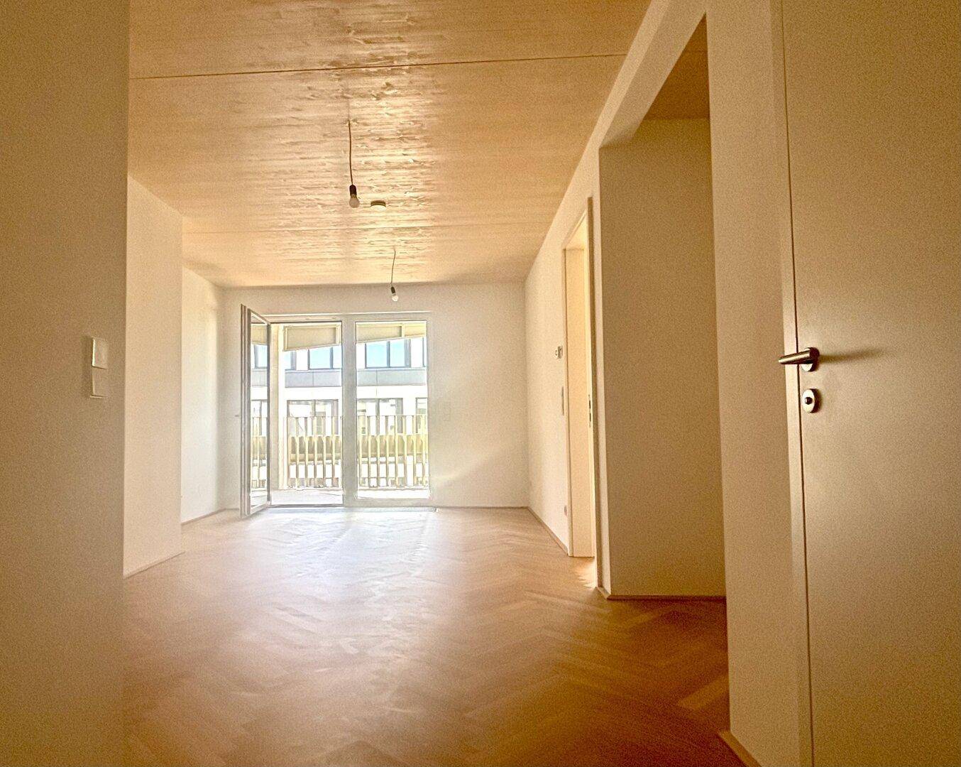 Heller Wohnraum mit Fischgrätparkett, großen Fenstern und Zugang zu einem Balkon.
