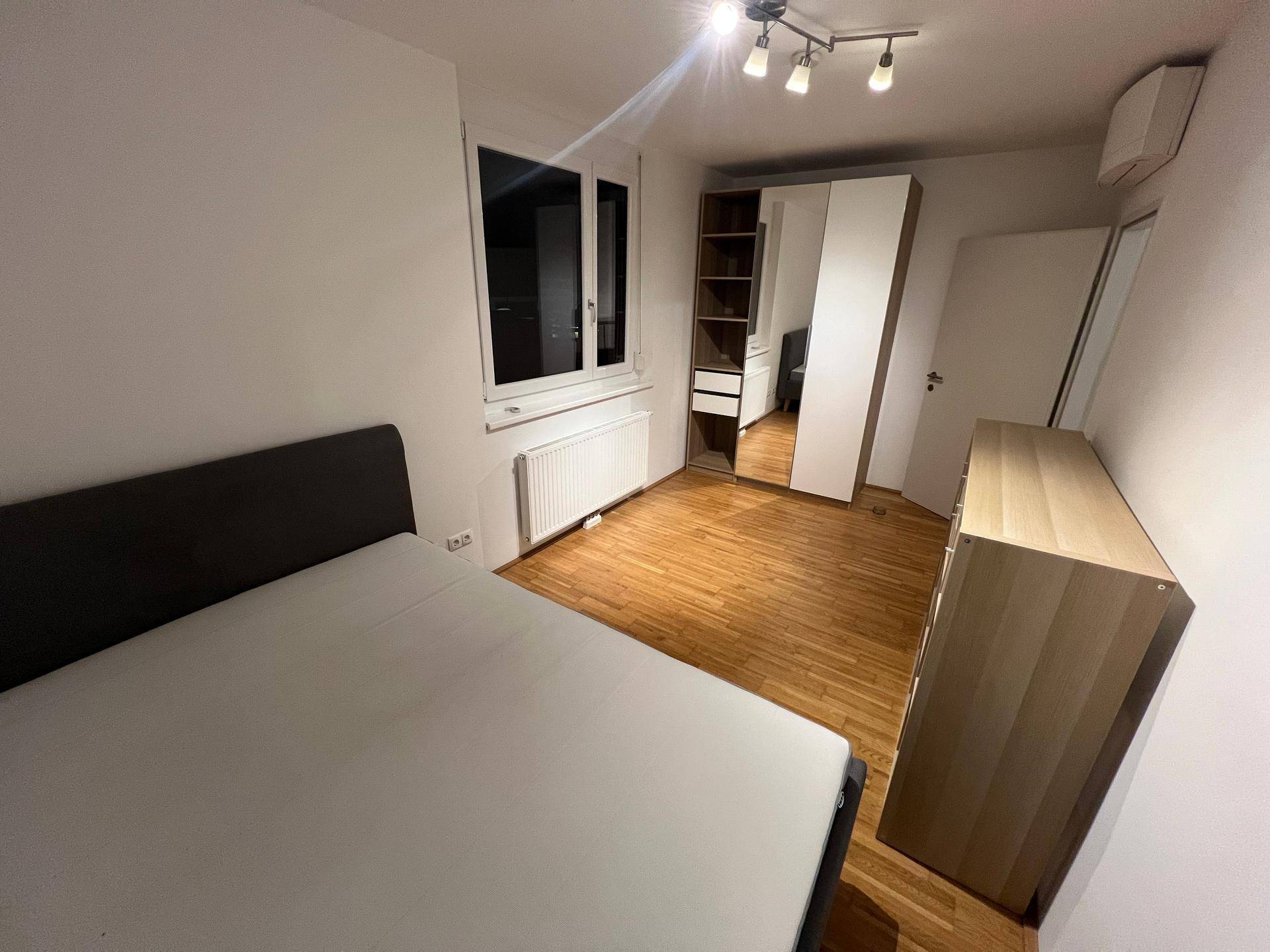 Funktionales Schlafzimmer mit hellem Parkettboden, großem Kleiderschrank und Fenster für natürliches Licht.