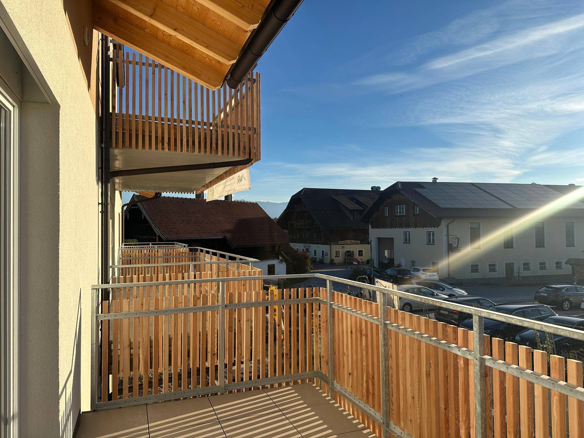 Balkon mit Holzzaun und Blick auf die Nachbarschaft sowie die umliegende Berglandschaft im Sonnenschein.