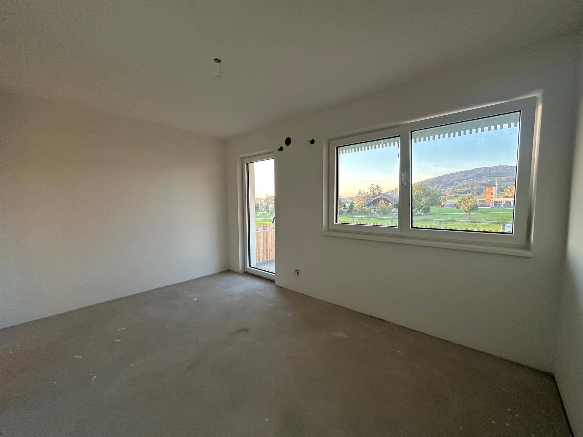 Unmöbliertes Schlafzimmer im Rohbau mit Balkonzugang und weitem Blick auf die umliegende Landschaft.