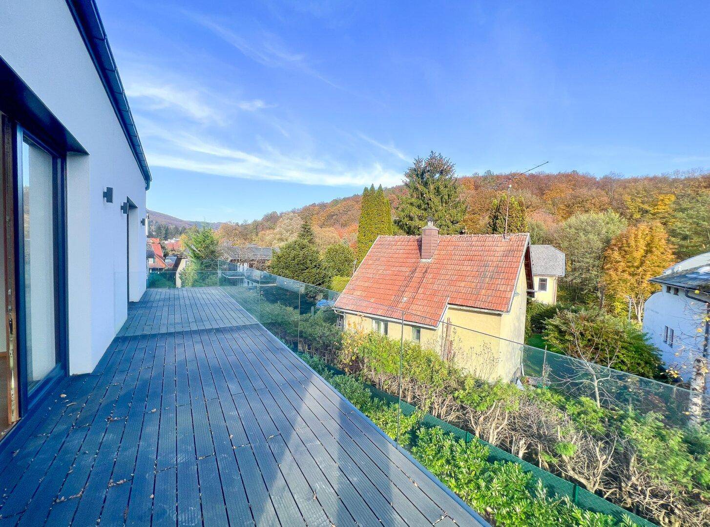 Großer Balkon mit dunklem Holzbelag und Glasgeländer, bietet einen schönen Blick ins Grüne.