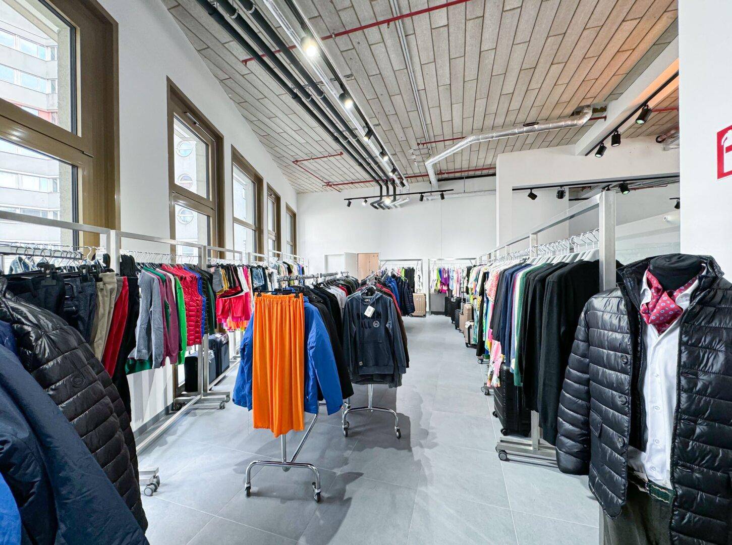 Weitläufiger Verkaufsraum mit Kleiderständern, großen Fenstern und industriellem Decken-Design.
