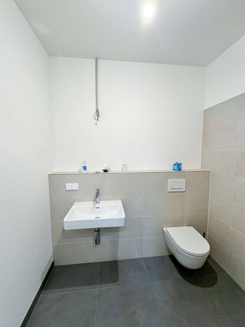 Schlichtes Badezimmer mit wandhängendem WC, Waschbecken und grauen Bodenfliesen.