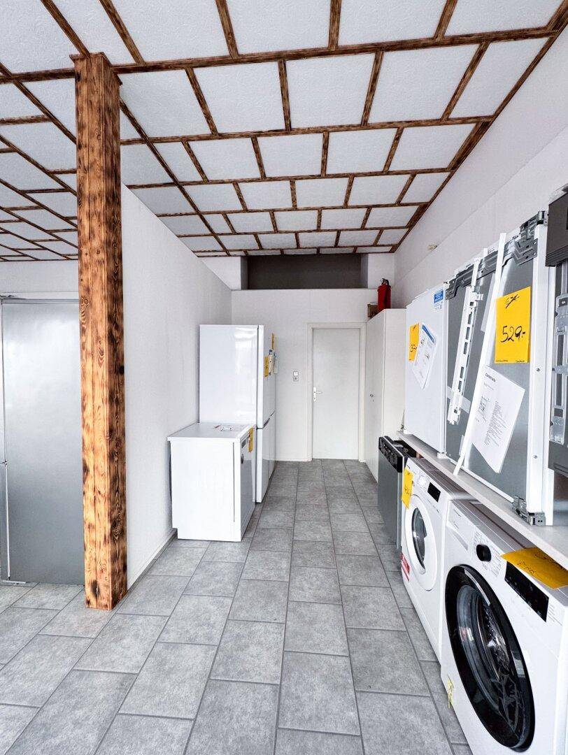 Langer Raum mit Fliesenboden, Holzsäule und einer Reihe von Haushaltsgeräten entlang der Wand.