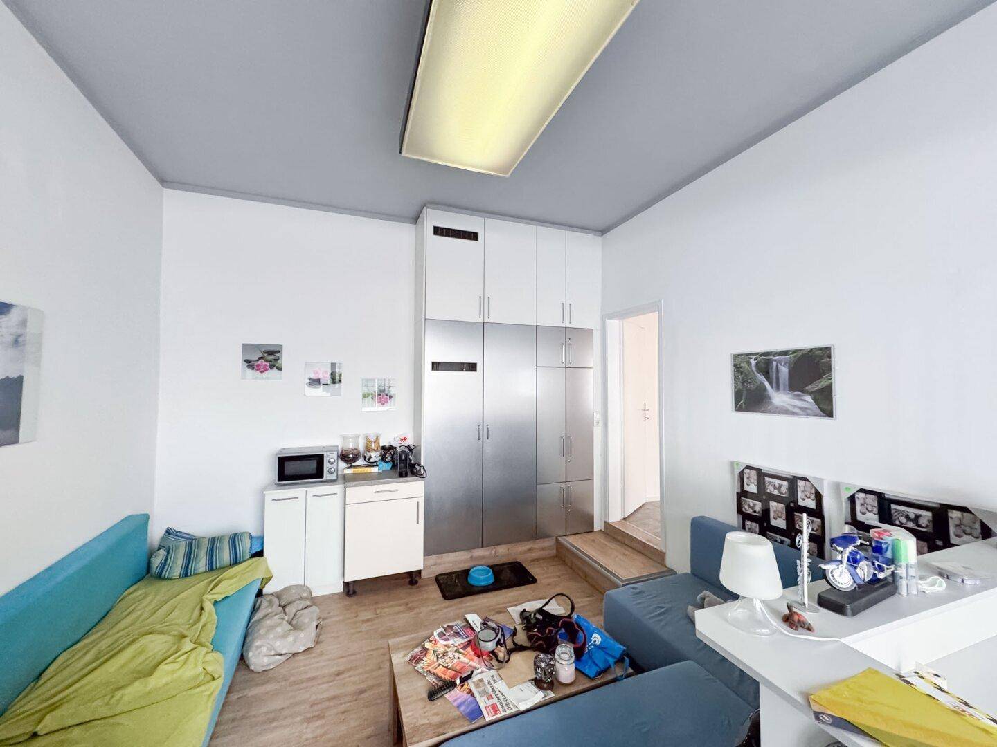 Gemütlicher Wohnbereich mit blauem Sofa, Laminatboden und einer kleinen Küchenzeile mit Einbauschränken.