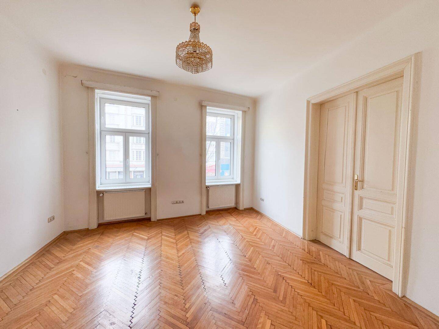 Heller Raum mit Fischgrätparkett, zwei Fenstern und einem eleganten Kristallkronleuchter.