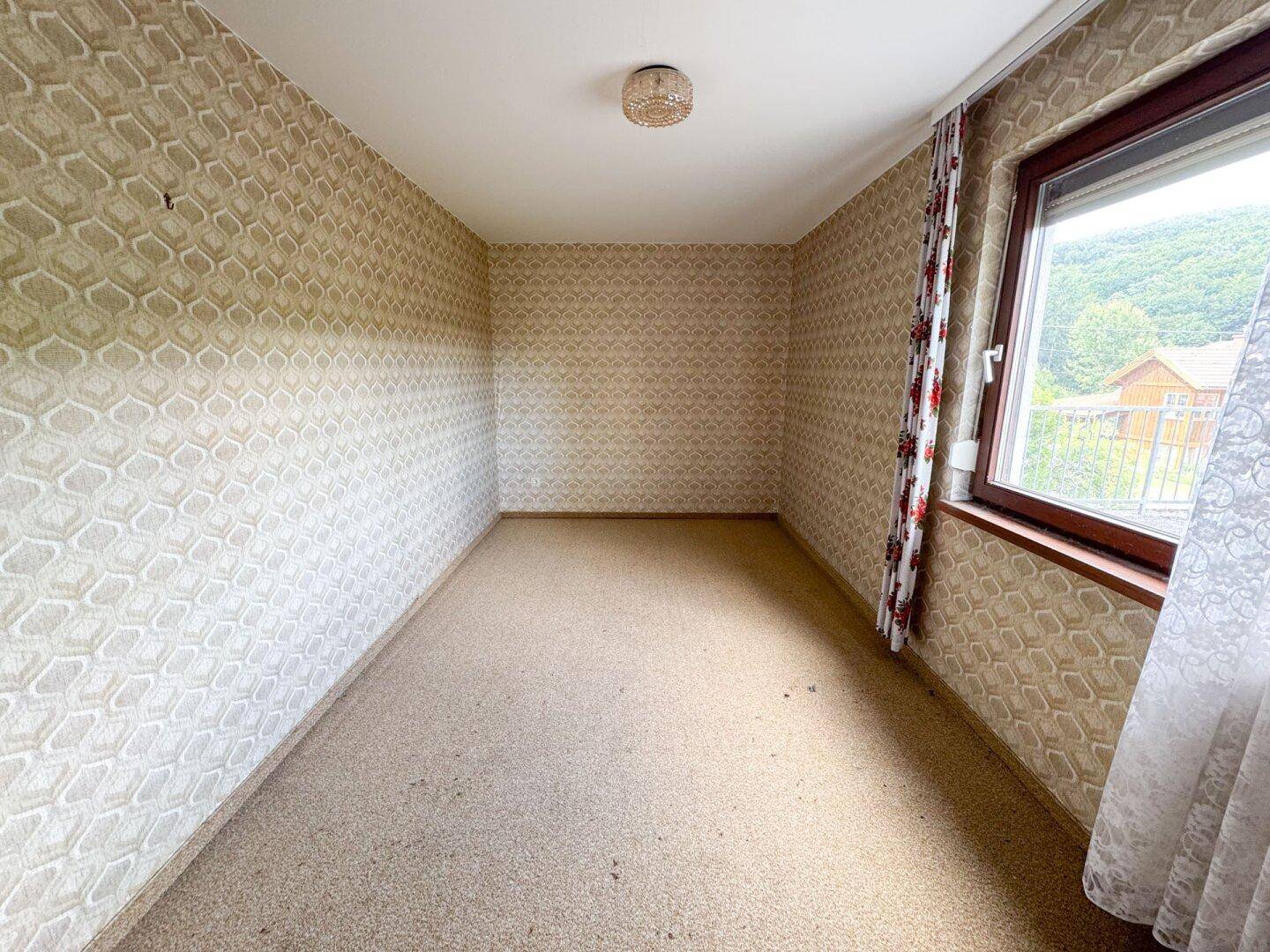 Gemütliches Zimmer mit gemusterter Tapete und Teppichboden, mit Blick ins Grüne.