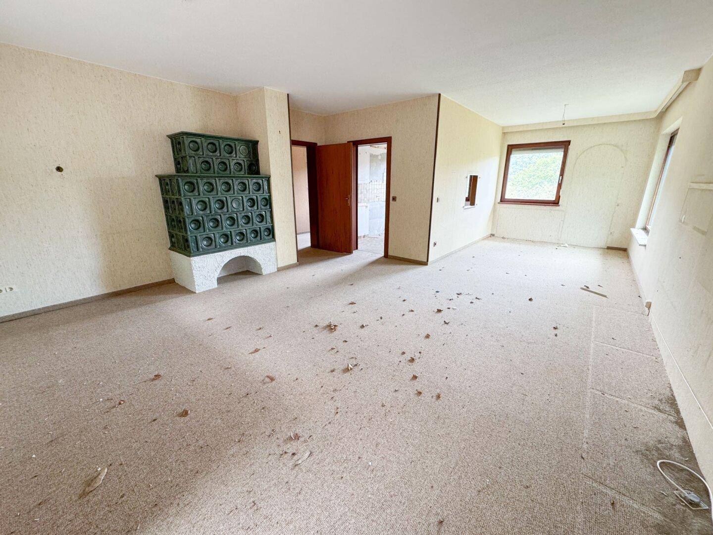 Geräumiges Wohnzimmer mit Kachelofen und Teppichboden, bietet viel Platz zur Gestaltung.