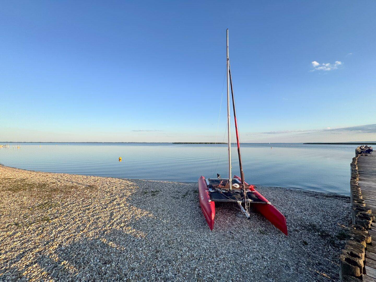 Roter Katamaran am Kiesstrand neben einem Holzsteg mit weitem Blick über das ruhige Wasser.