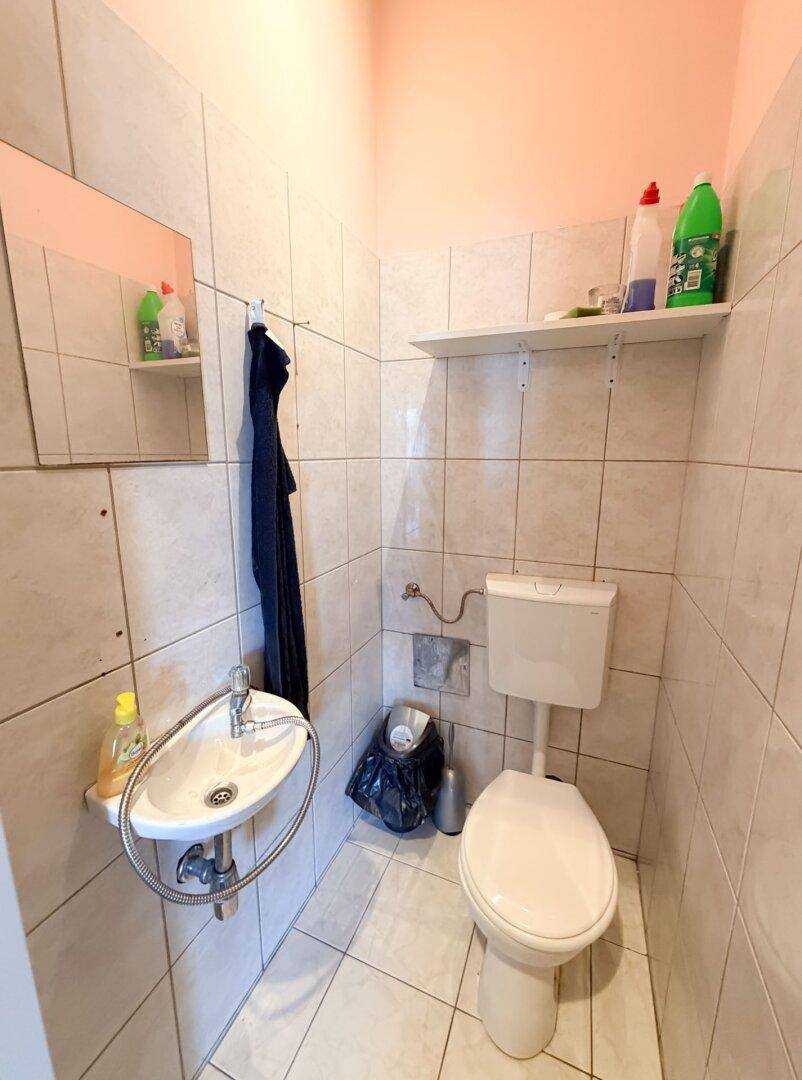 Einfache Toilette mit Waschbecken, Spiegel und Fliesenboden, funktional und sauber gehalten.