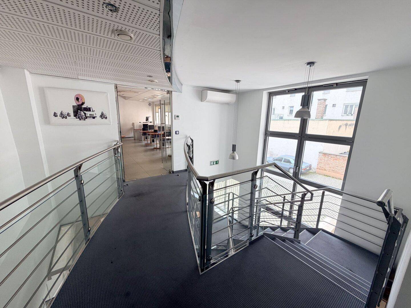 Treppenhaus mit Metallgeländer, Teppichboden und Blick in einen angrenzenden Büroraum.