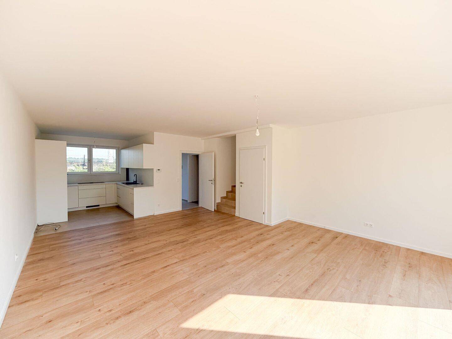 Heller offener Wohnbereich mit Holzboden, integrierter Küchenzeile und Zugang zu einer Treppe.