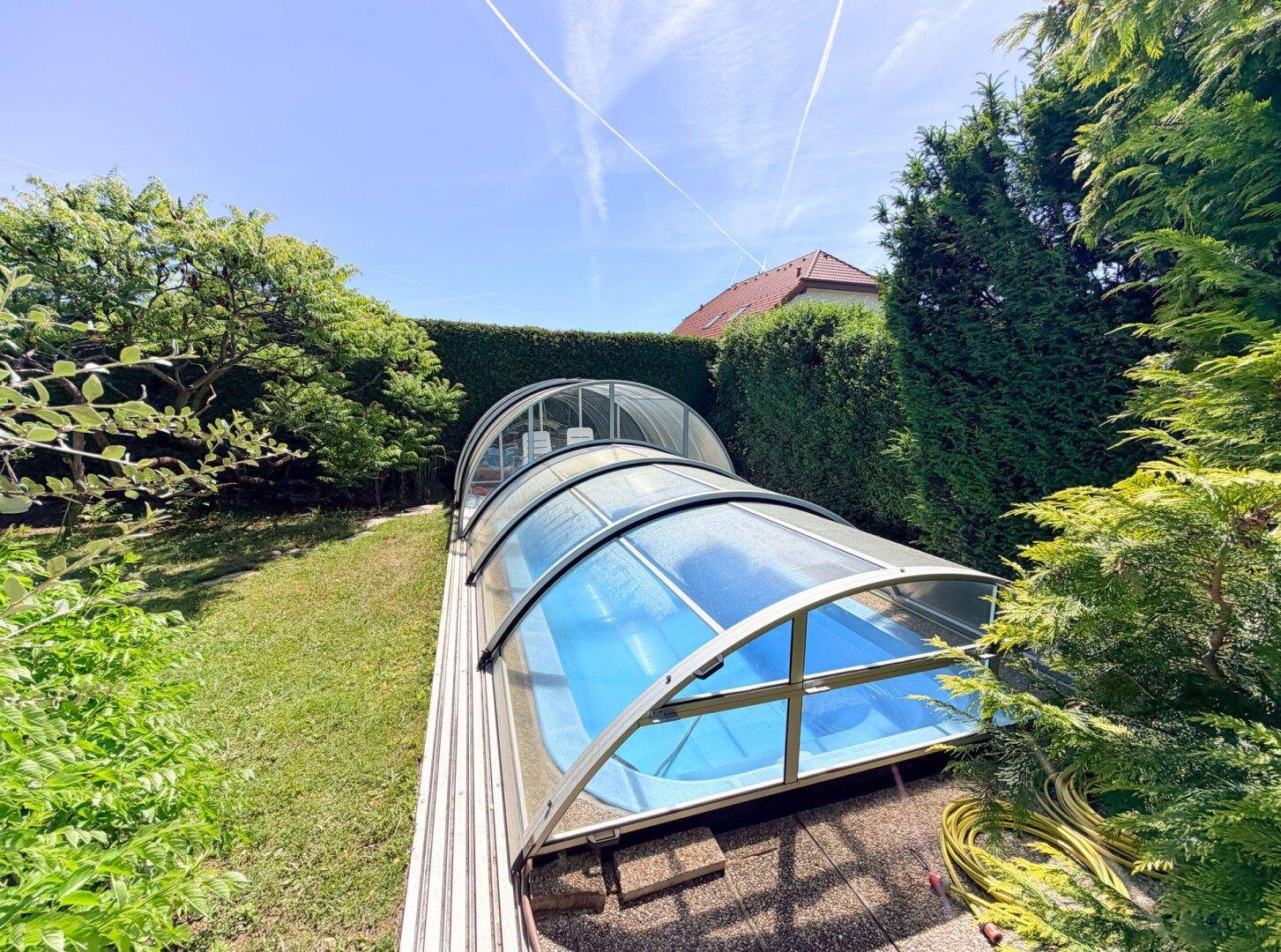 Überdachter Swimmingpool im Garten, umgeben von hohen Hecken und Bäumen an einem sonnigen Tag.